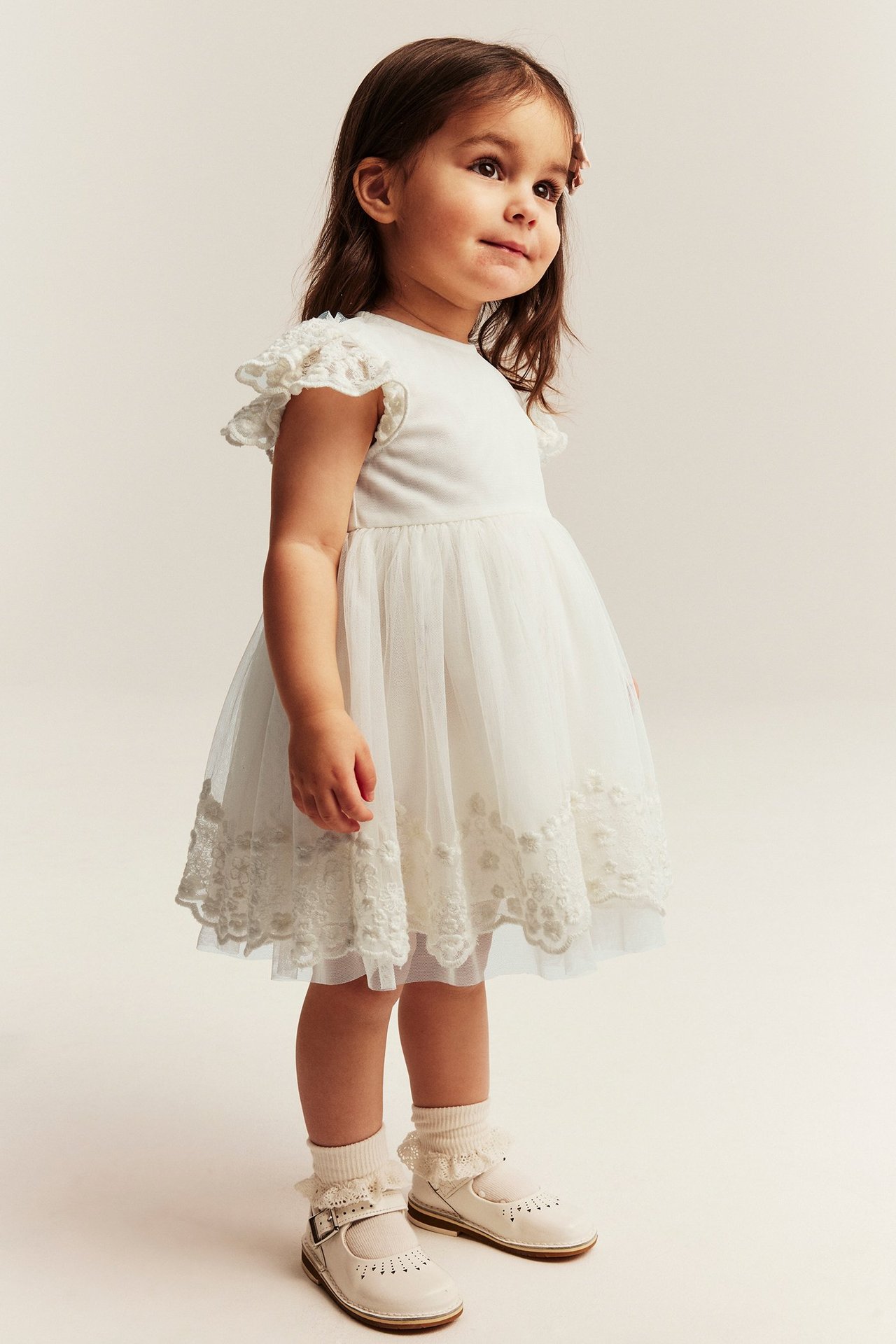 Sidovy, liten tjej i offwhite broderad meshklänning med volangaxlar, volangstrumpor och vita Mary Jane-skor.