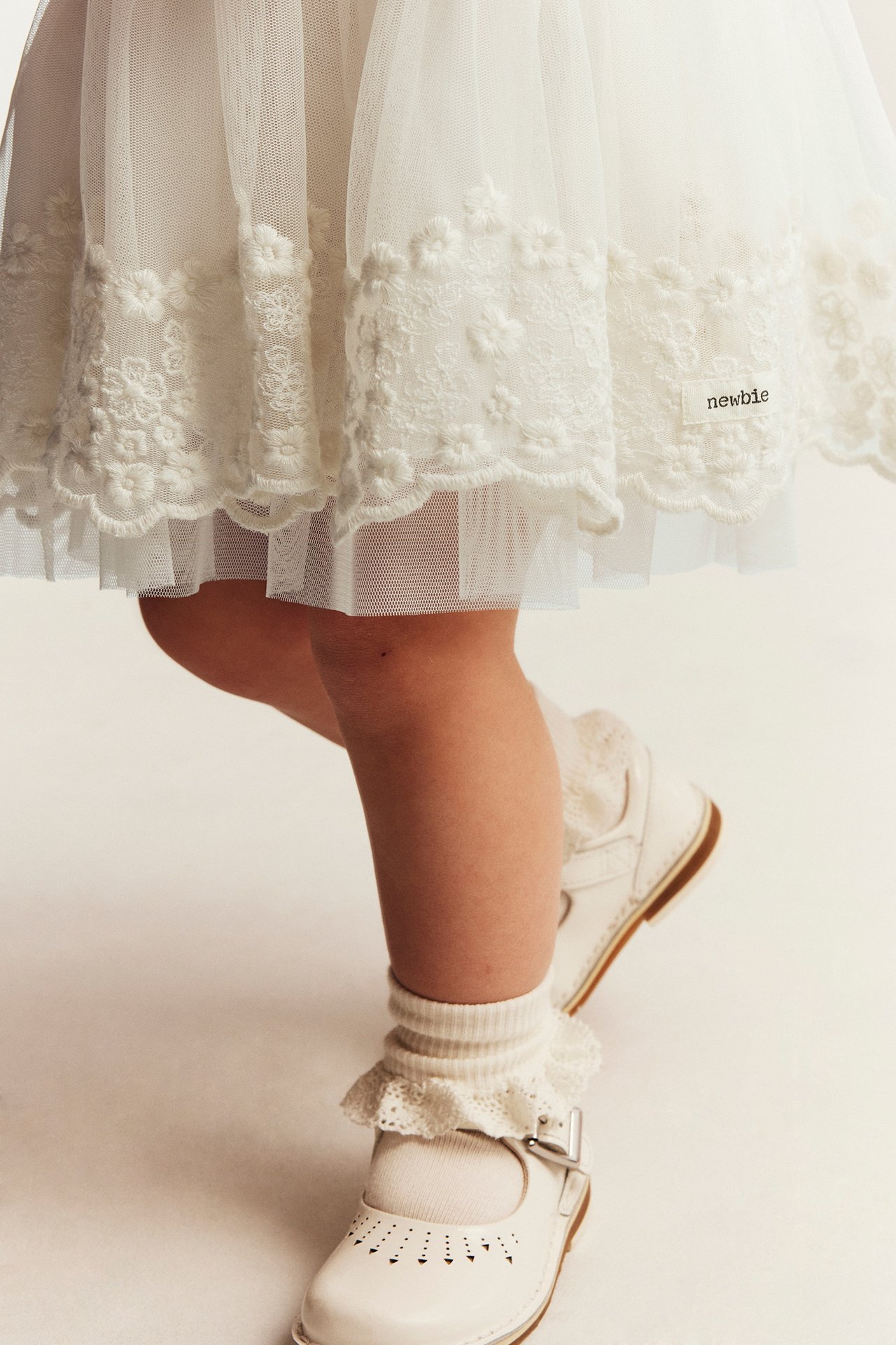 Zbliżenie: off-white siatkowa sukienka dziecięca z haftem, białe skarpetki z falbanką i buty Mary Jane.