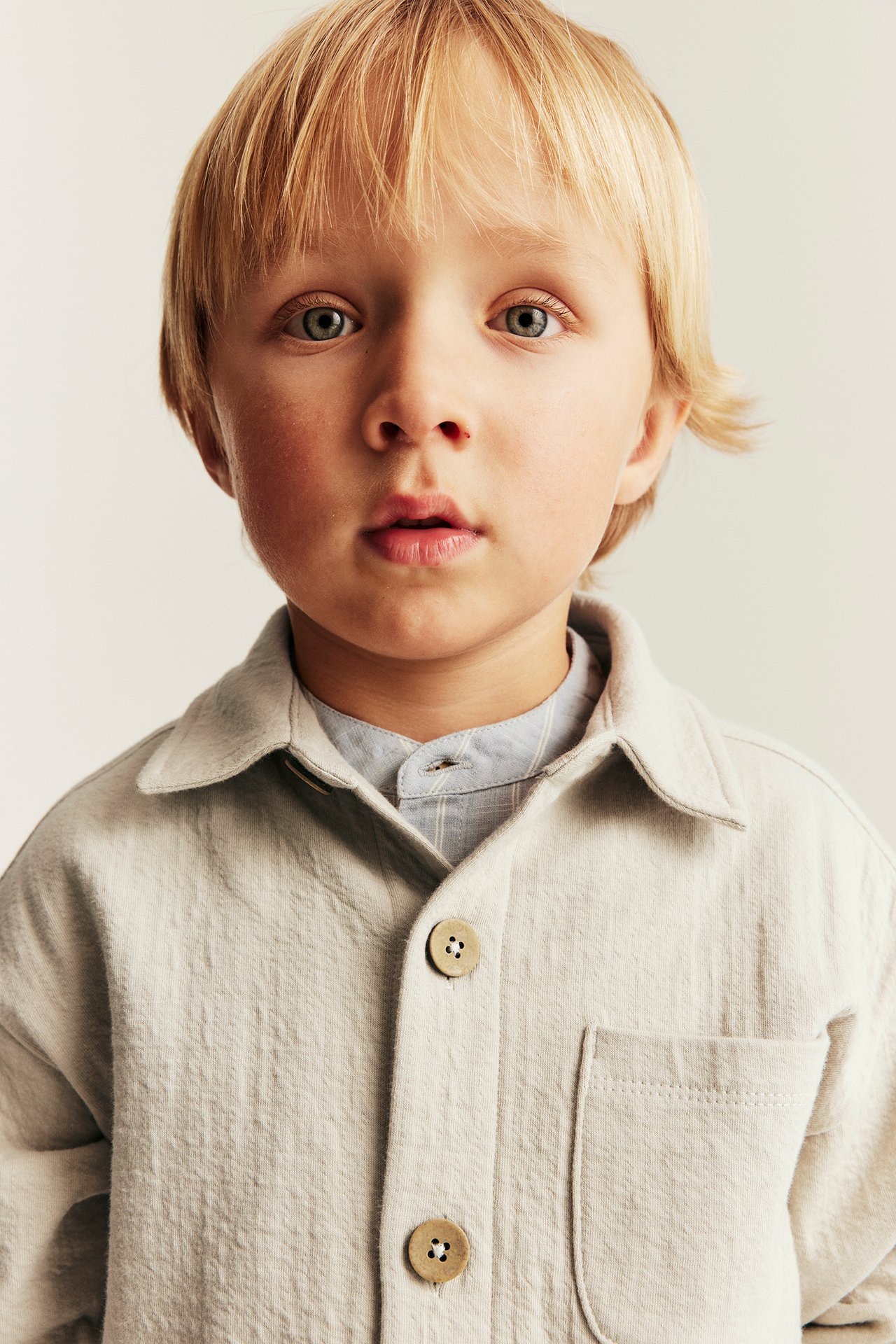 Nærbilde forfra av barn i beige teksturert overshirt med knapper over lyseblå stripet skjorte.