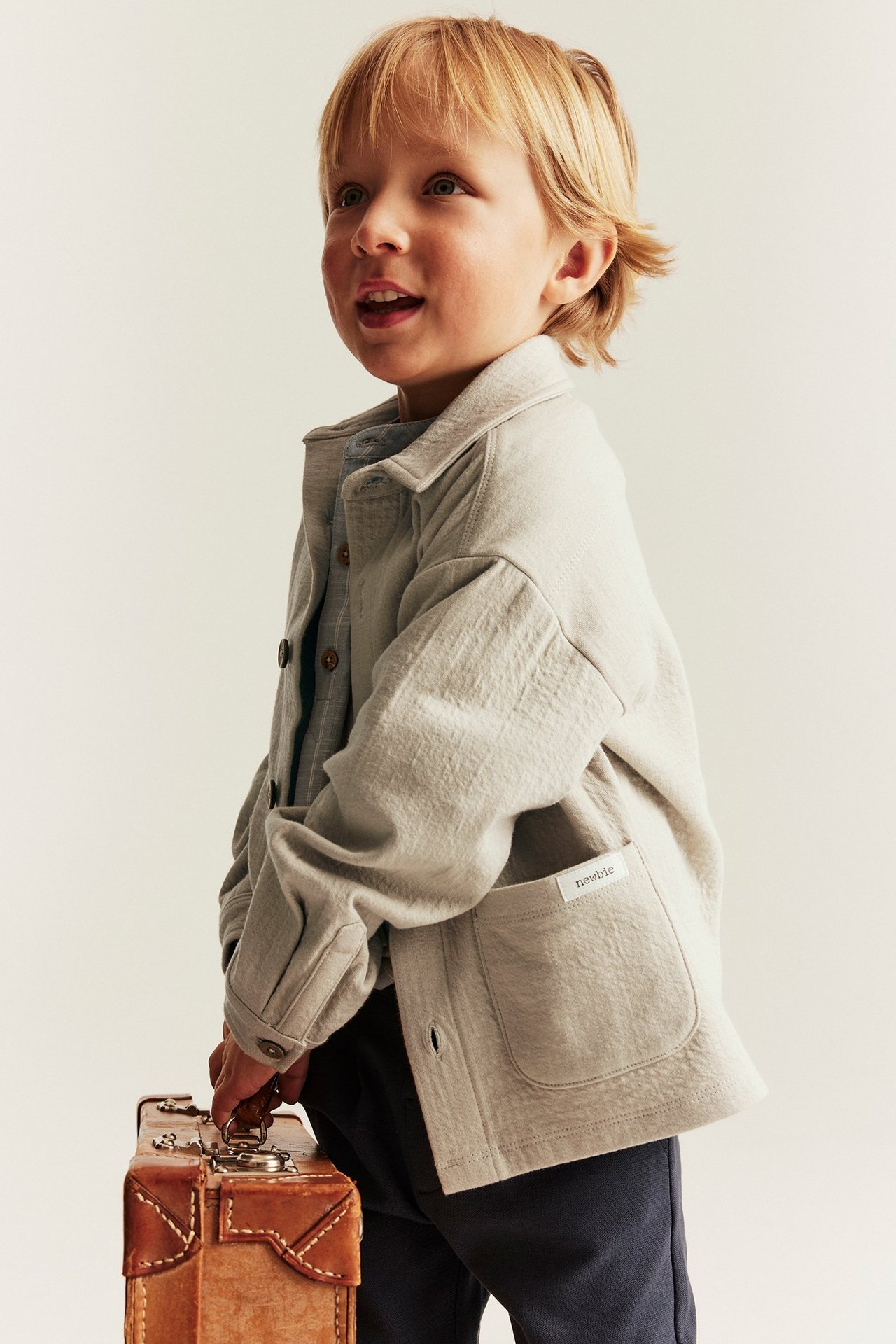 Barn i beigefarget overskjorte med krage over blå skjorte, mørke bukser, holder en brun koffert.