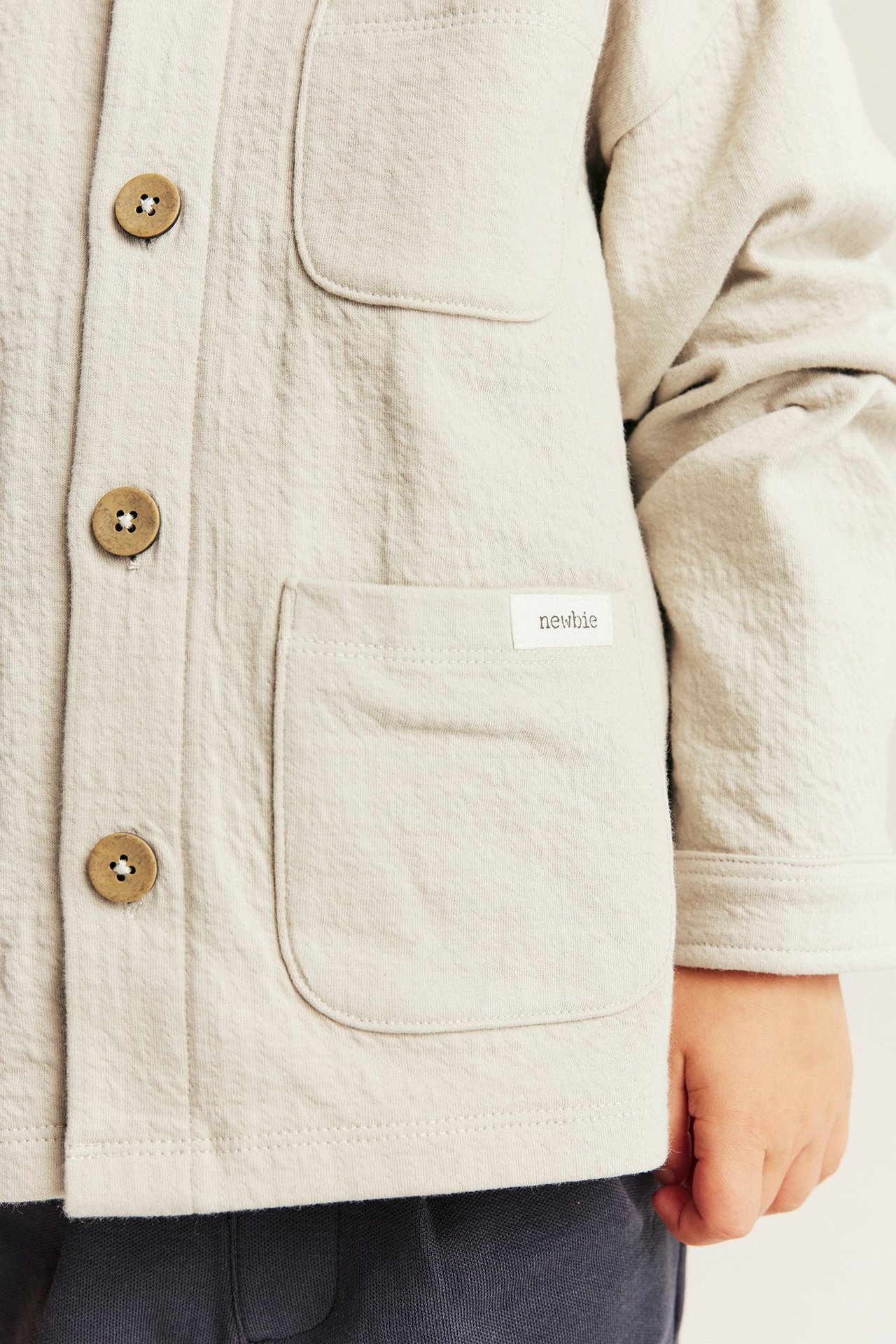 Nærbilde av et barn i beige strukturstrikket overskjorte med krage, treknapper og frontlommer.