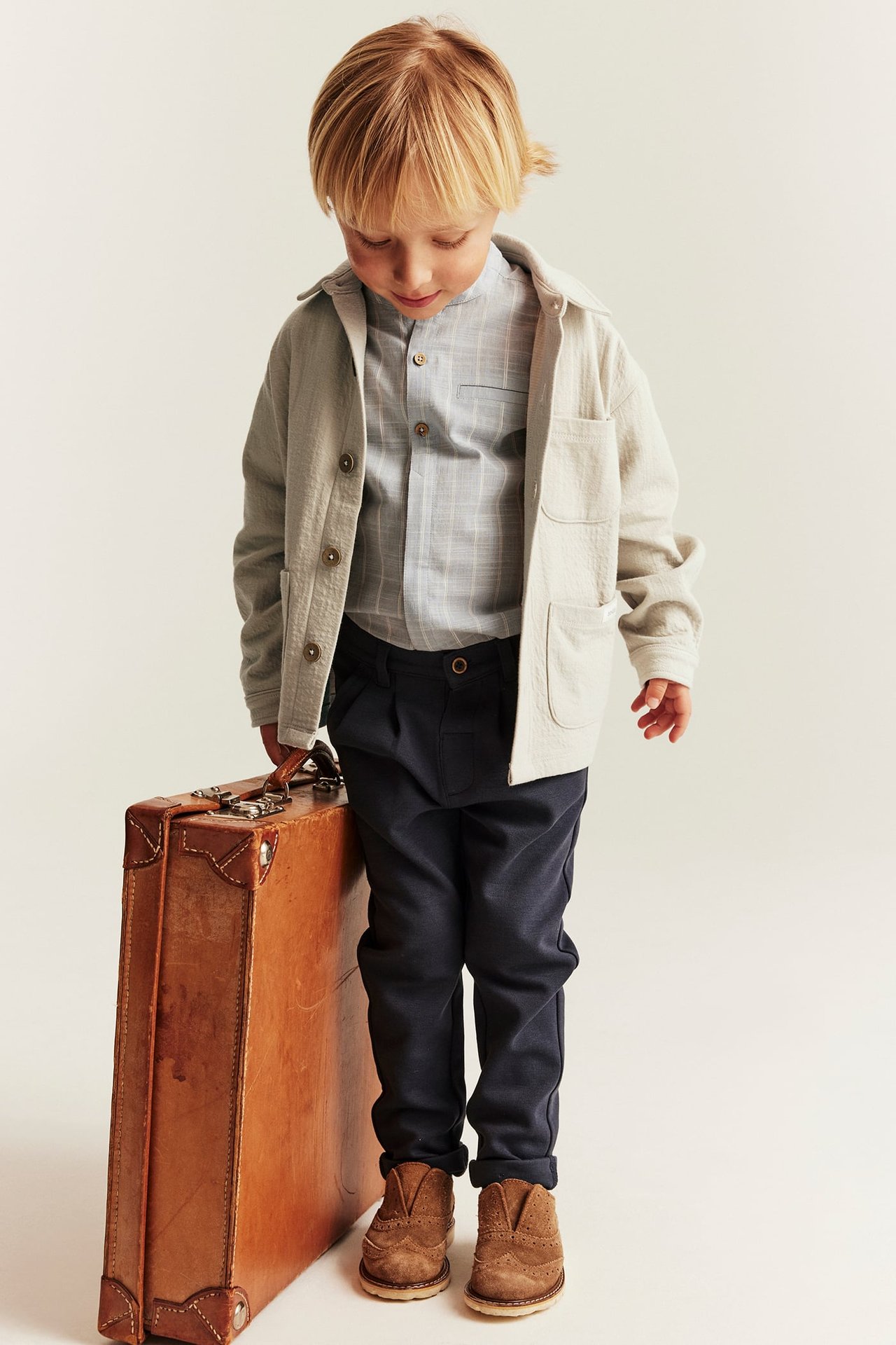 Barn i beige strukturerad overshirt, rutig skjorta, mörka byxor, bruna skor, bredvid resväska.