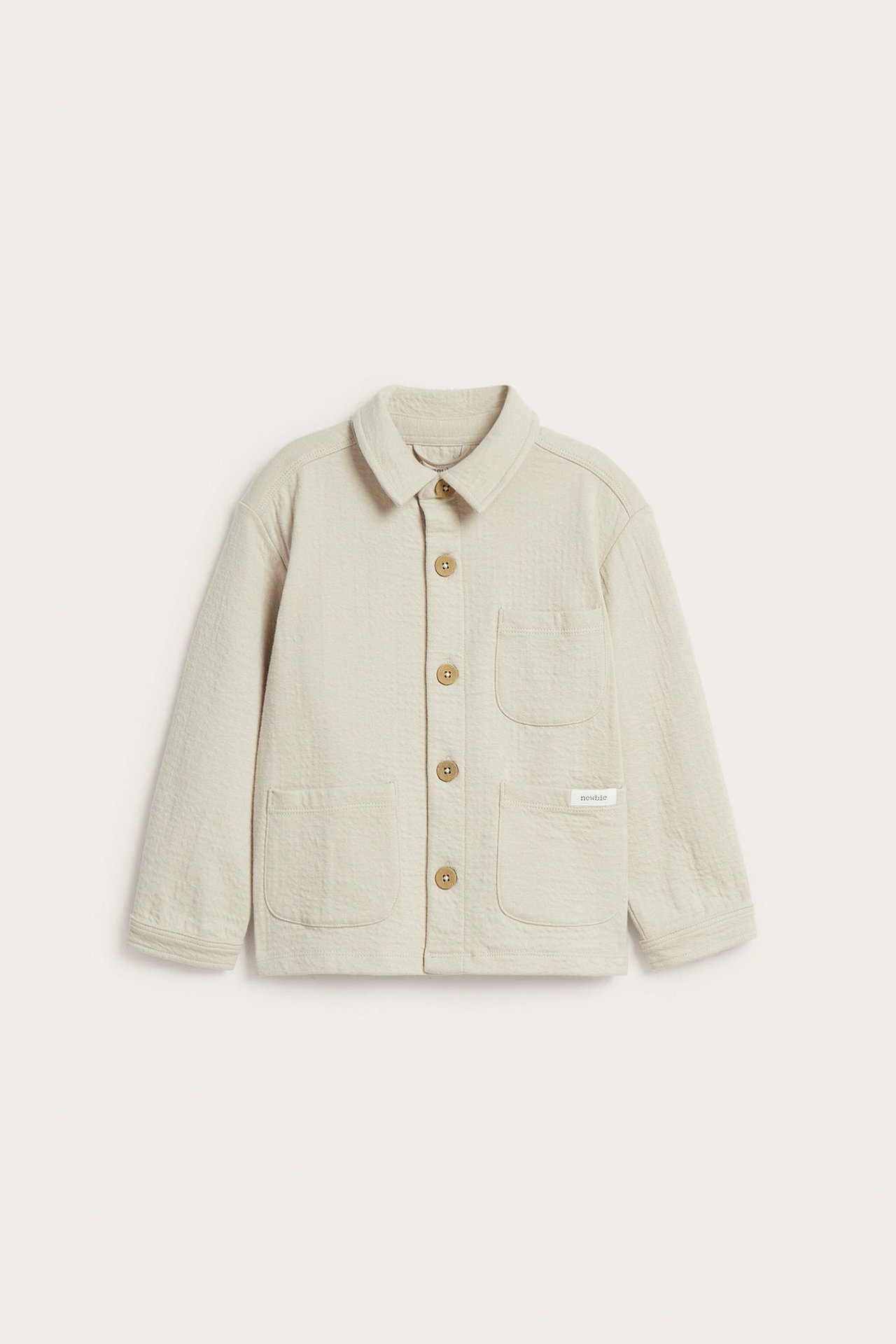 Framsida av en beige overshirt i strukturtrikå för barn med krage, fem knappar och tre fickor.