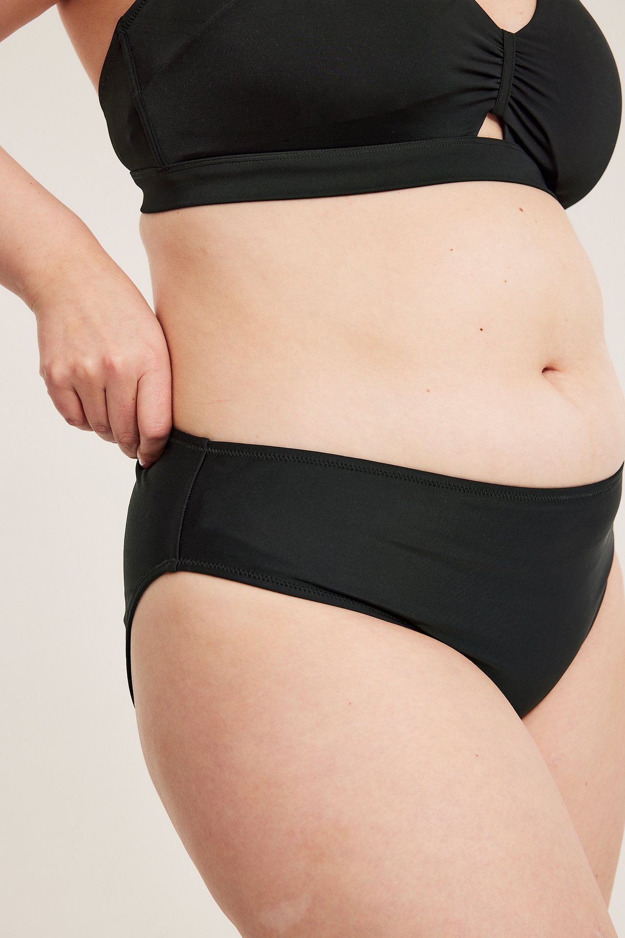 Sidebilde av kvinne i ensfarget svart bikiniunderdel, hånden justerer den, med svart bikinitopp.