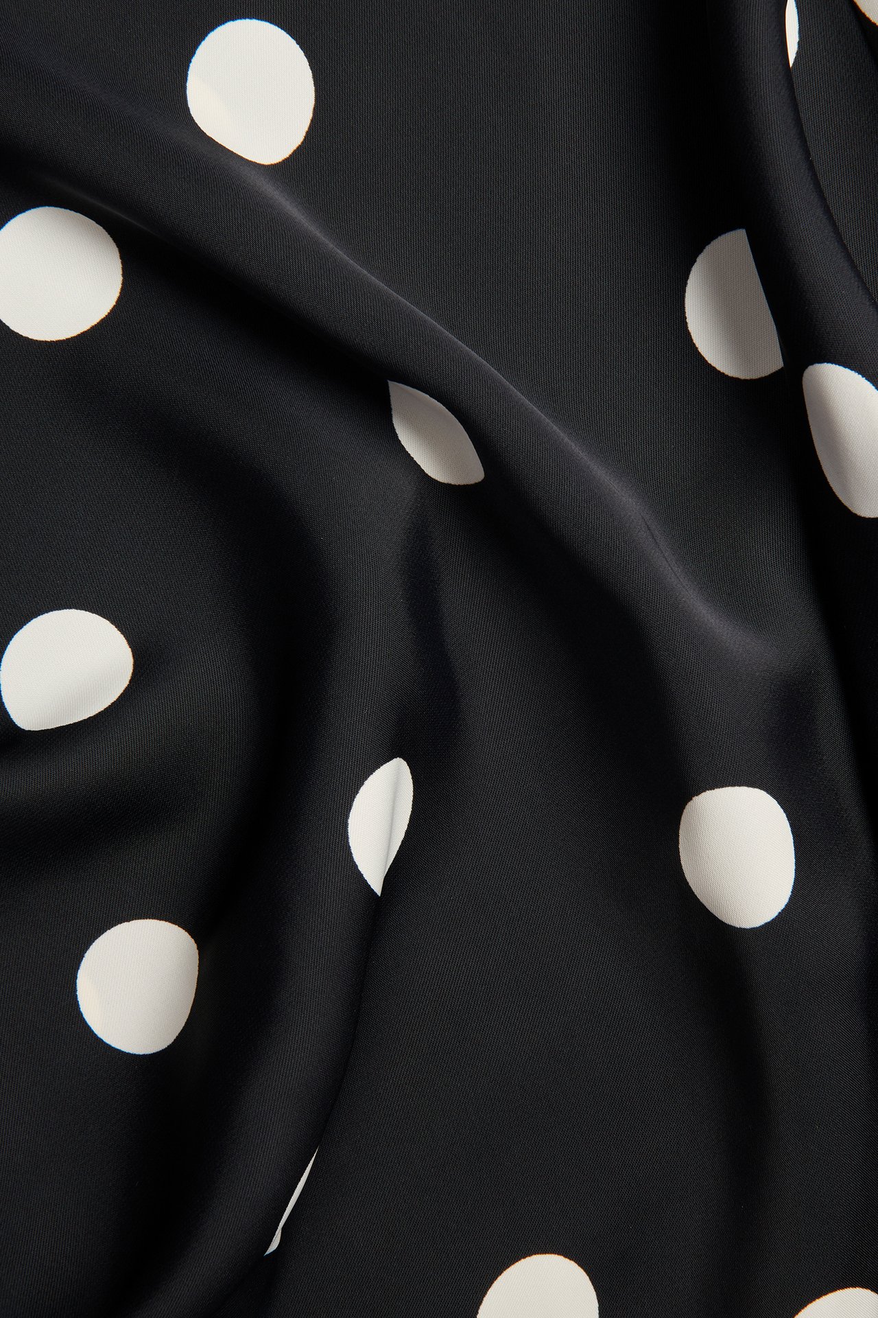 Nærbilde av svart glansfullt polyesterskjørt med hvitt prikket mønster.