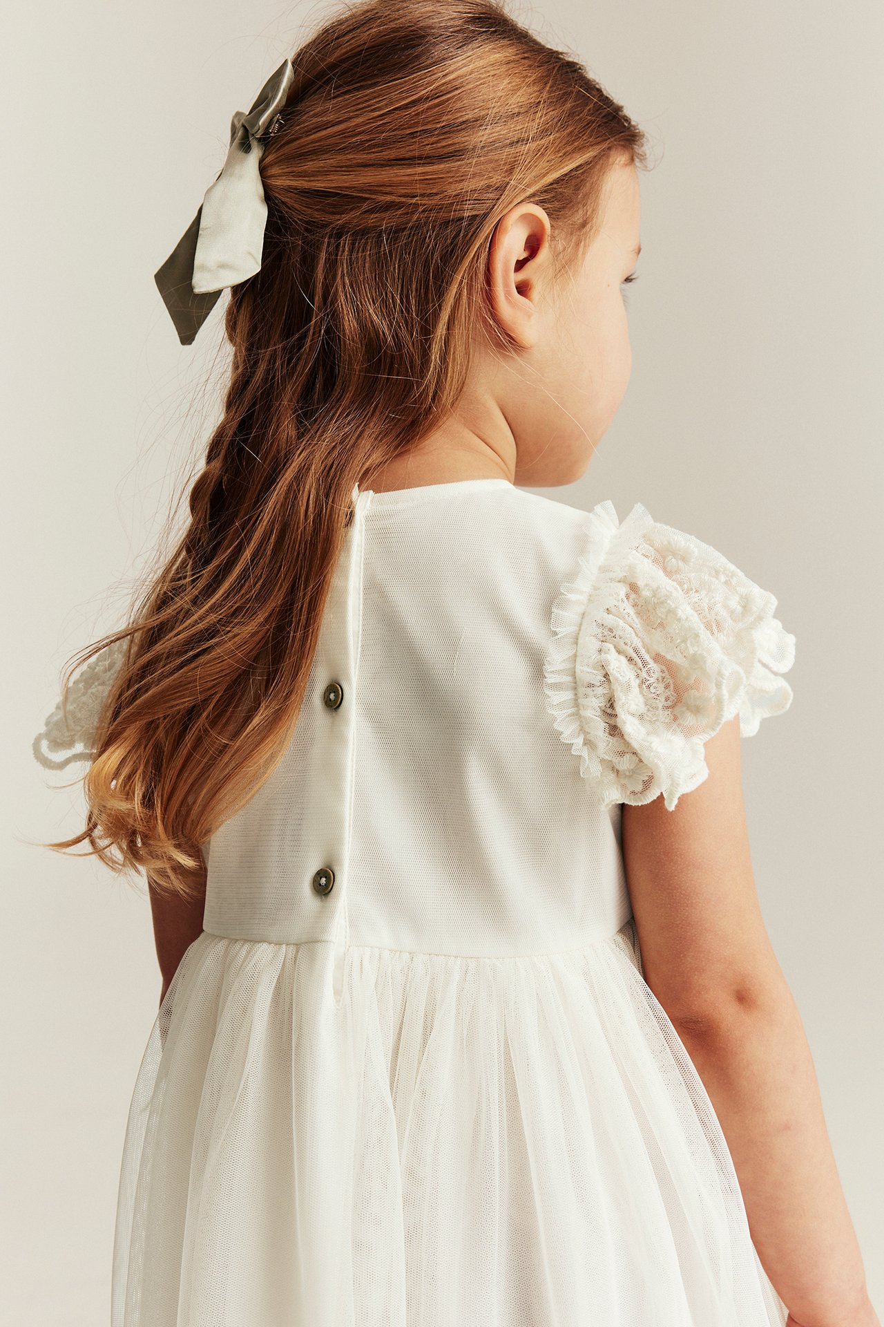 Ryggvy av ett barn i offwhite meshklänning med broderade volanger, knappar bak och hårrosett.