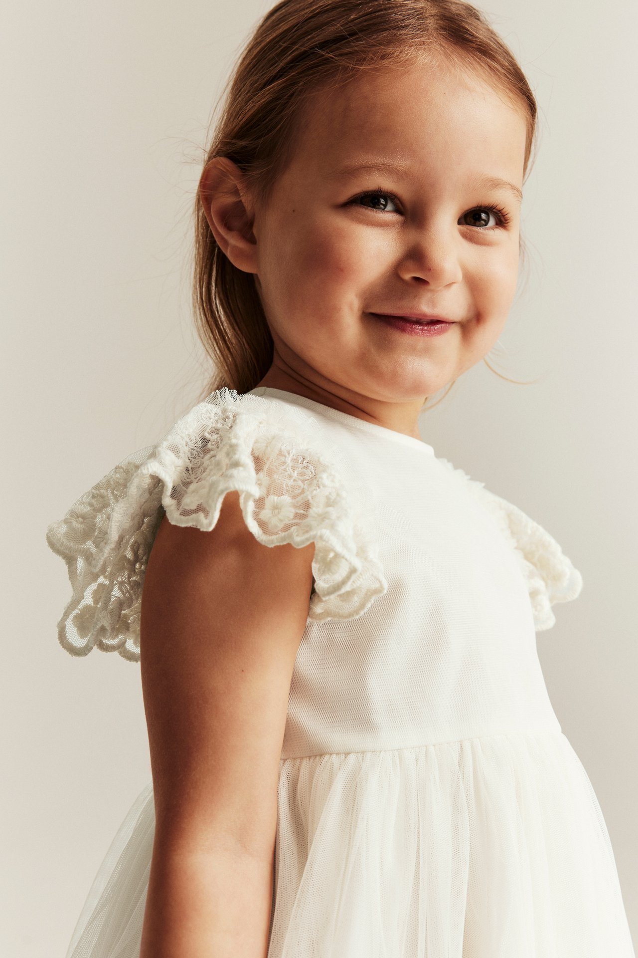 Närbild framifrån på ett barn i en offwhite meshklänning med broderade spetsvolanger på axlarna.