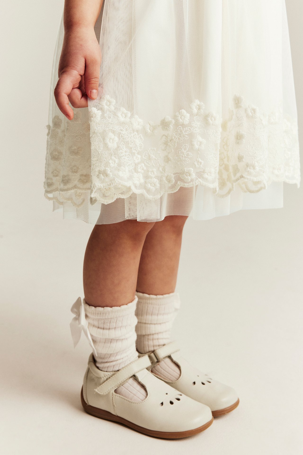 Närbild av ett barn i en offwhite broderad meshklänning, ribbstickade strumpor med rosetter och Mary Jane-skor.