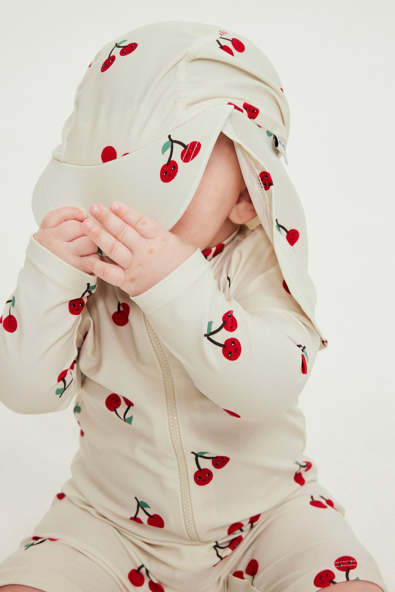 Närbild på en baby i ljusbeige UV-keps med körsbärsmönster, matchande en långärmad UV-dräkt.
