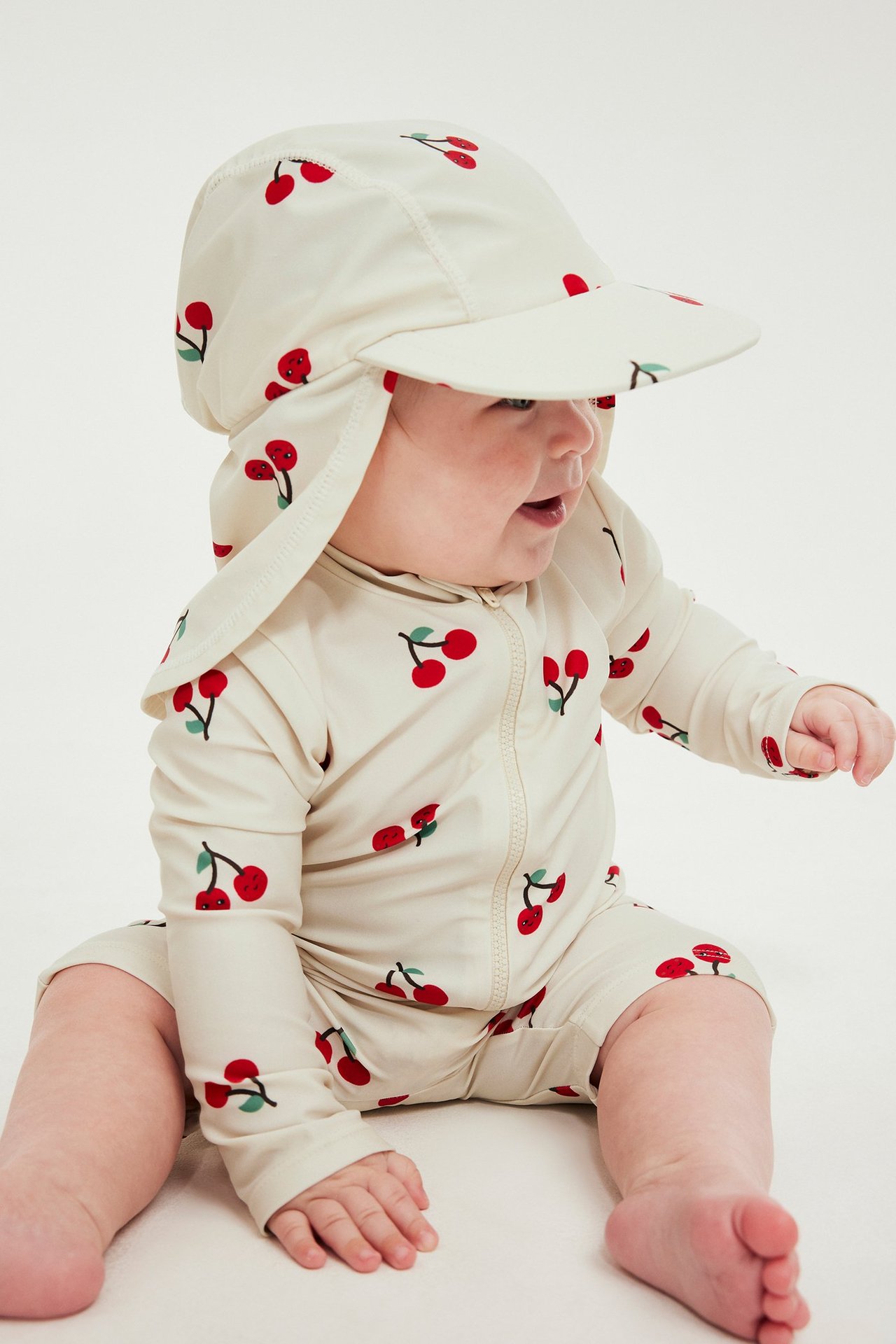 Baby i en ljusbeige UV-keps med körsbärsmönster och nackskydd, samt matchande långärmad UV-baddräkt.