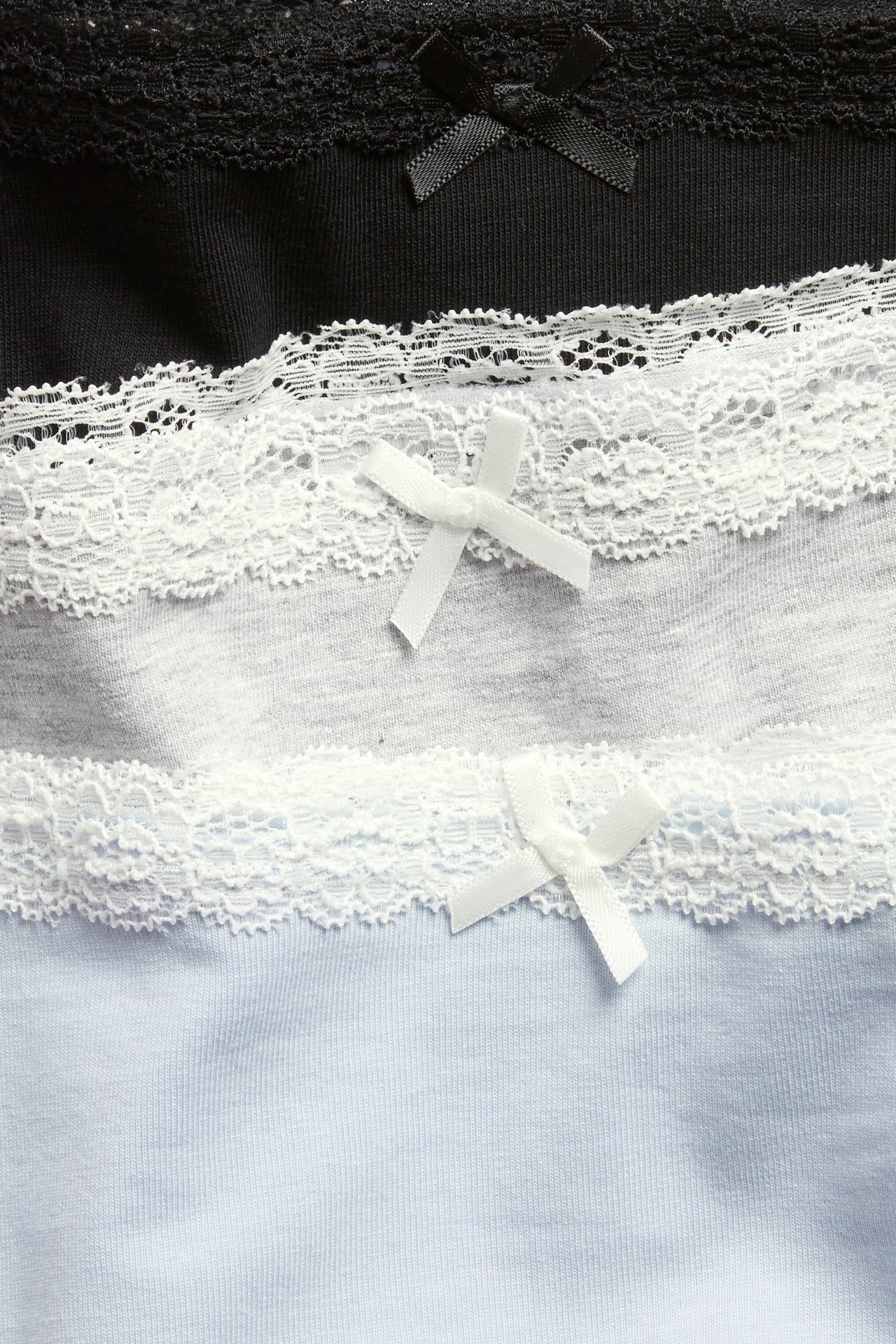 Zbliżenie: trzy bawełniane bokserki w czerni, szarości, jasnym błękicie, z koronką i kokardkami.