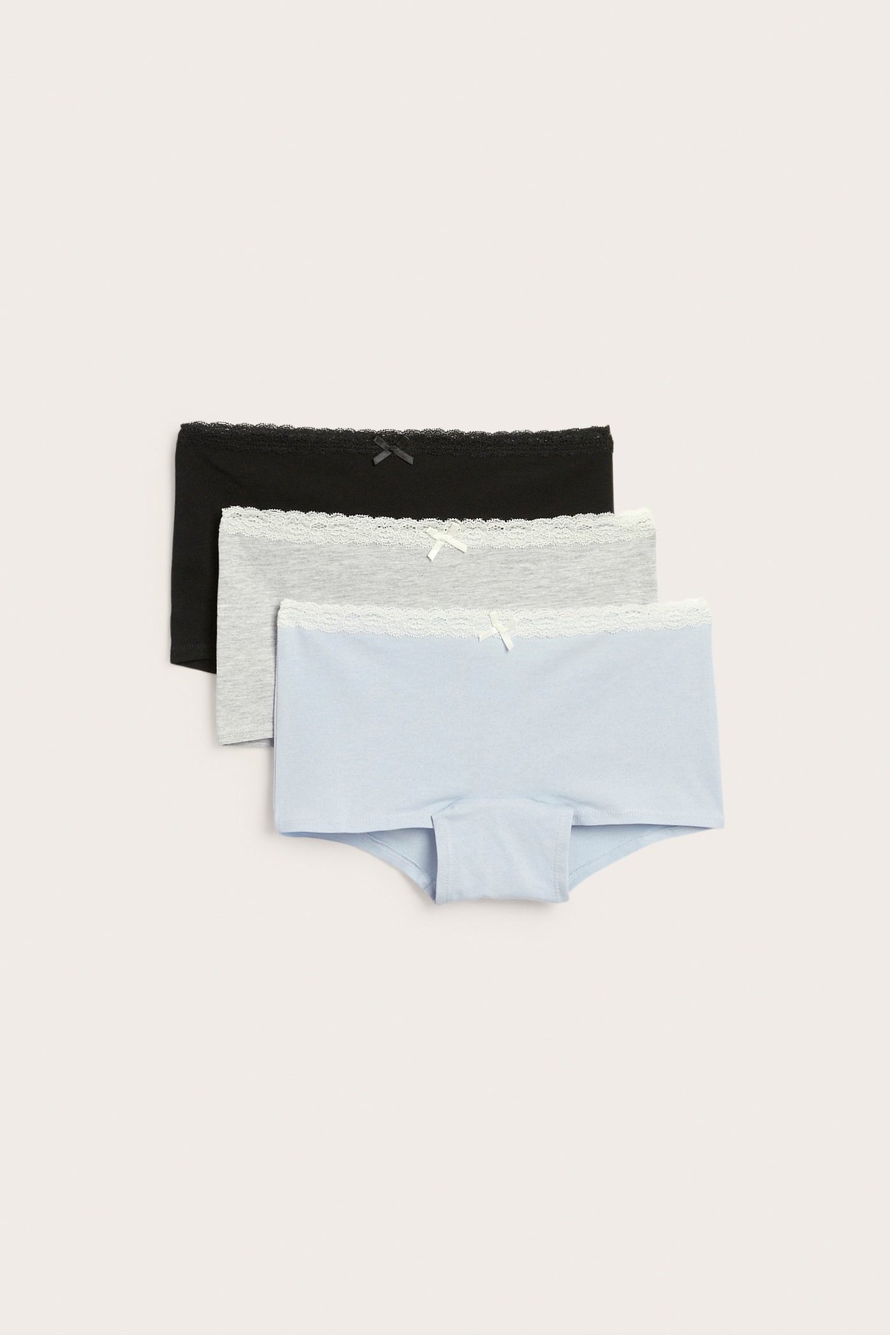 Widok z przodu trzech bokserek: czarnych, szarych i jasnoniebieskich, wszystkie z białą koronką i kokardkami.