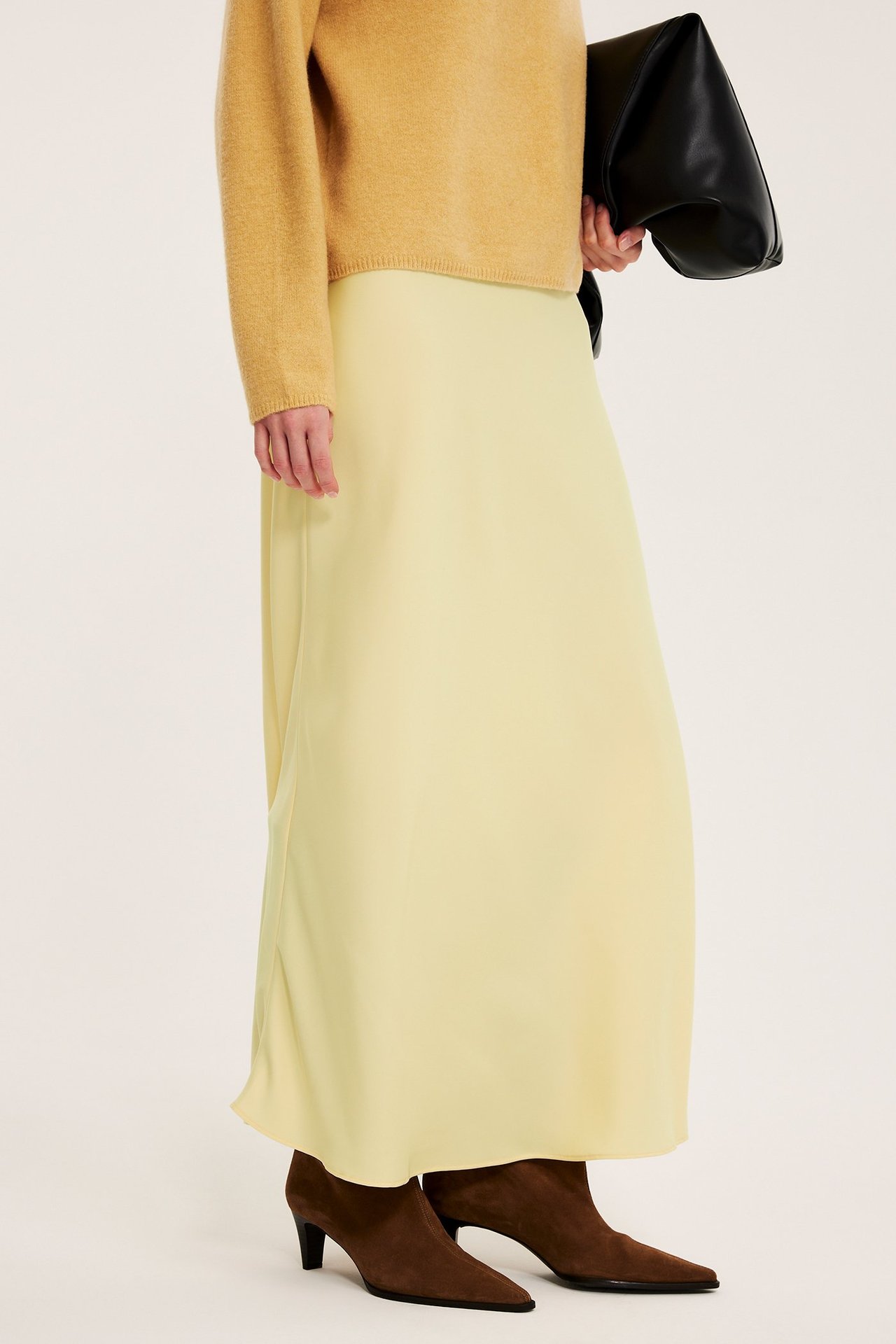 Naisen sivukuva vaaleankeltaisessa satiinihameessa, vaaleanruskeassa neulepuserossa, mustassa käsilaukussa ja ruskeissa nilkkureissa.