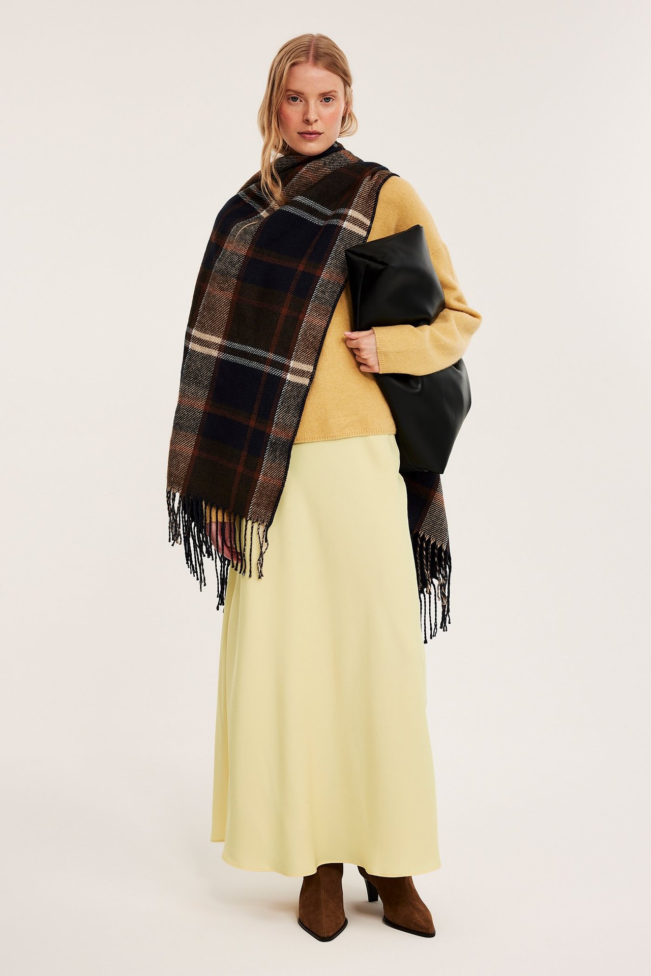 Nainen yllään vaaleankeltainen satiinihame, keltainen neule, ruudullinen huivi, ruskeat nilkkurit, musta clutch.