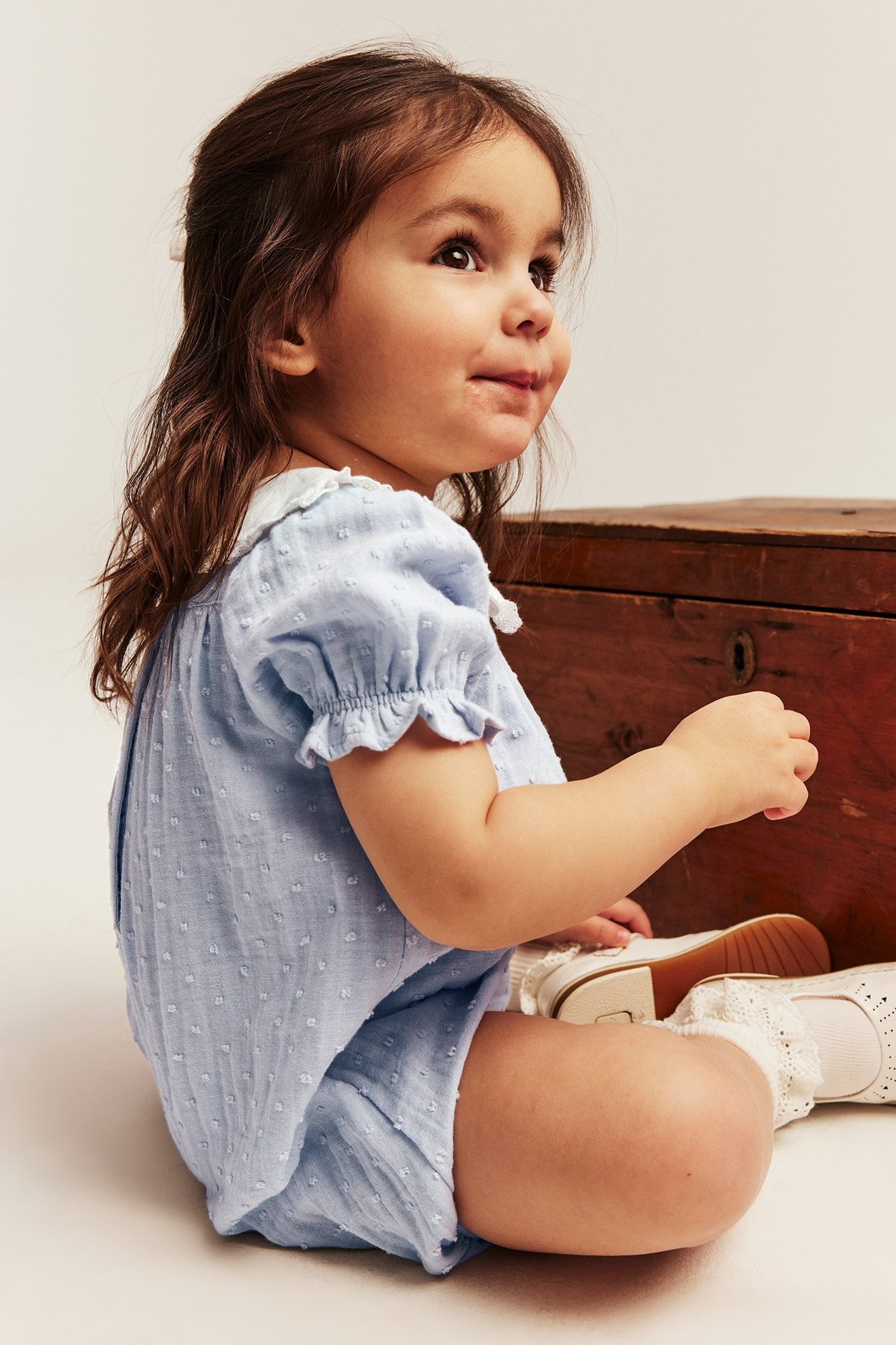 Sidovy av bebis i ljusblå prickig playsuit med vit sjömanskrage, vita strumpor och skor.