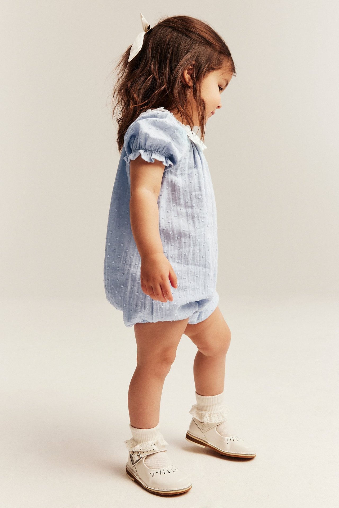Sidevisning: baby i lys blå prikkete playsuit med hvit sjømannskrage, hvite sokker og lyse beige sko.