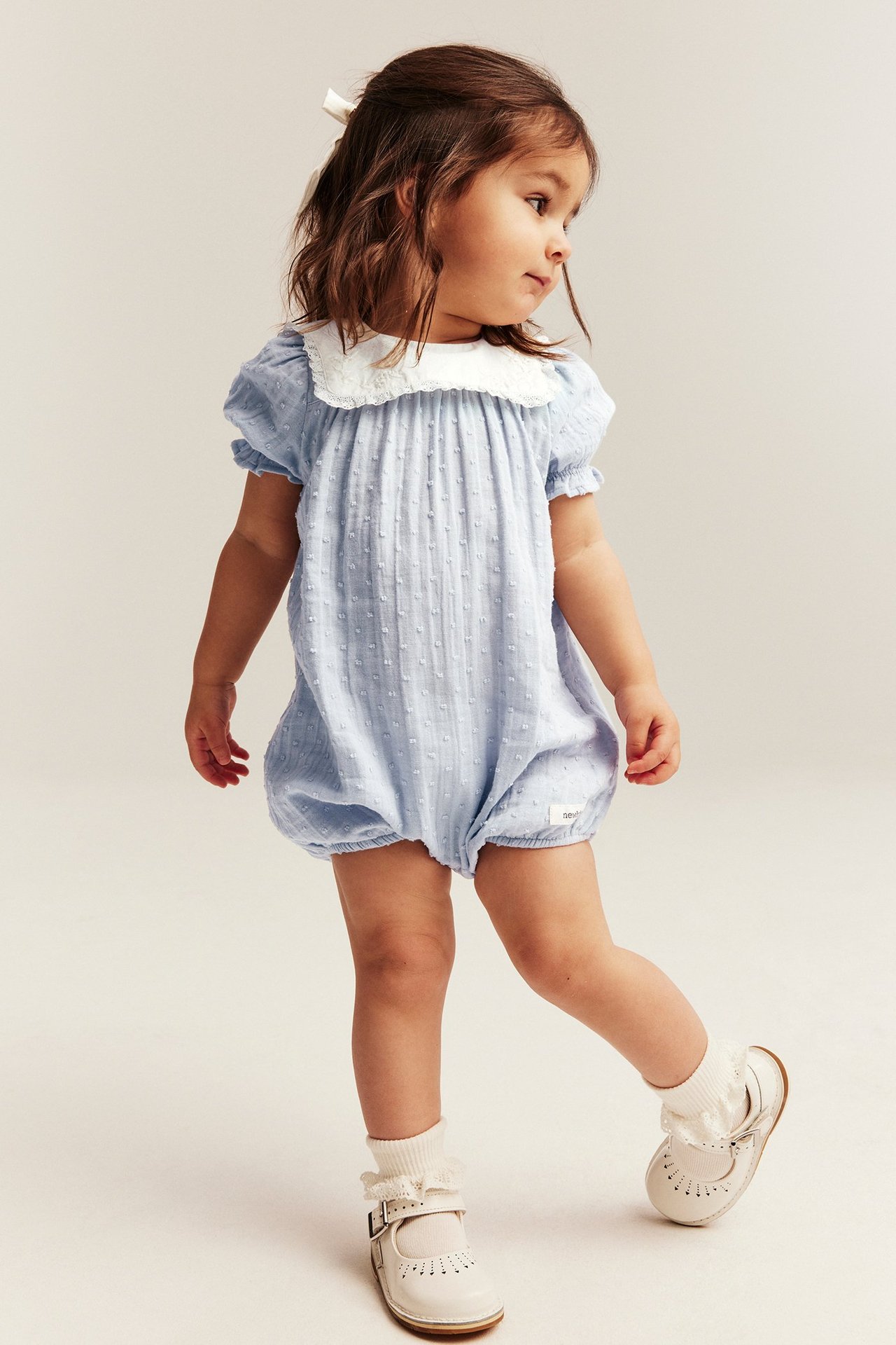 Forfra: baby i lyseblå prikket playsuit med hvit blondekrage og hvite Mary Jane-sko.