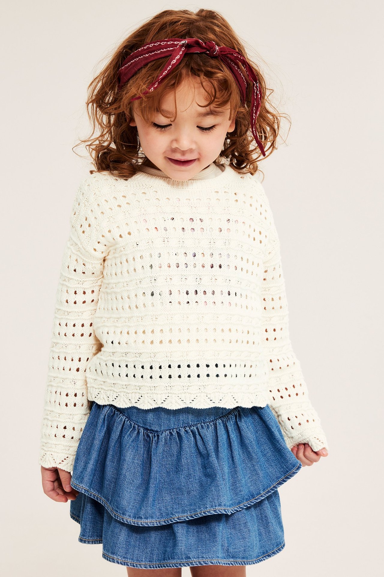 Framifrån: Barn i offwhite hålstickad bomullströja, blå jeanskjol med volanger och röd bandana.
