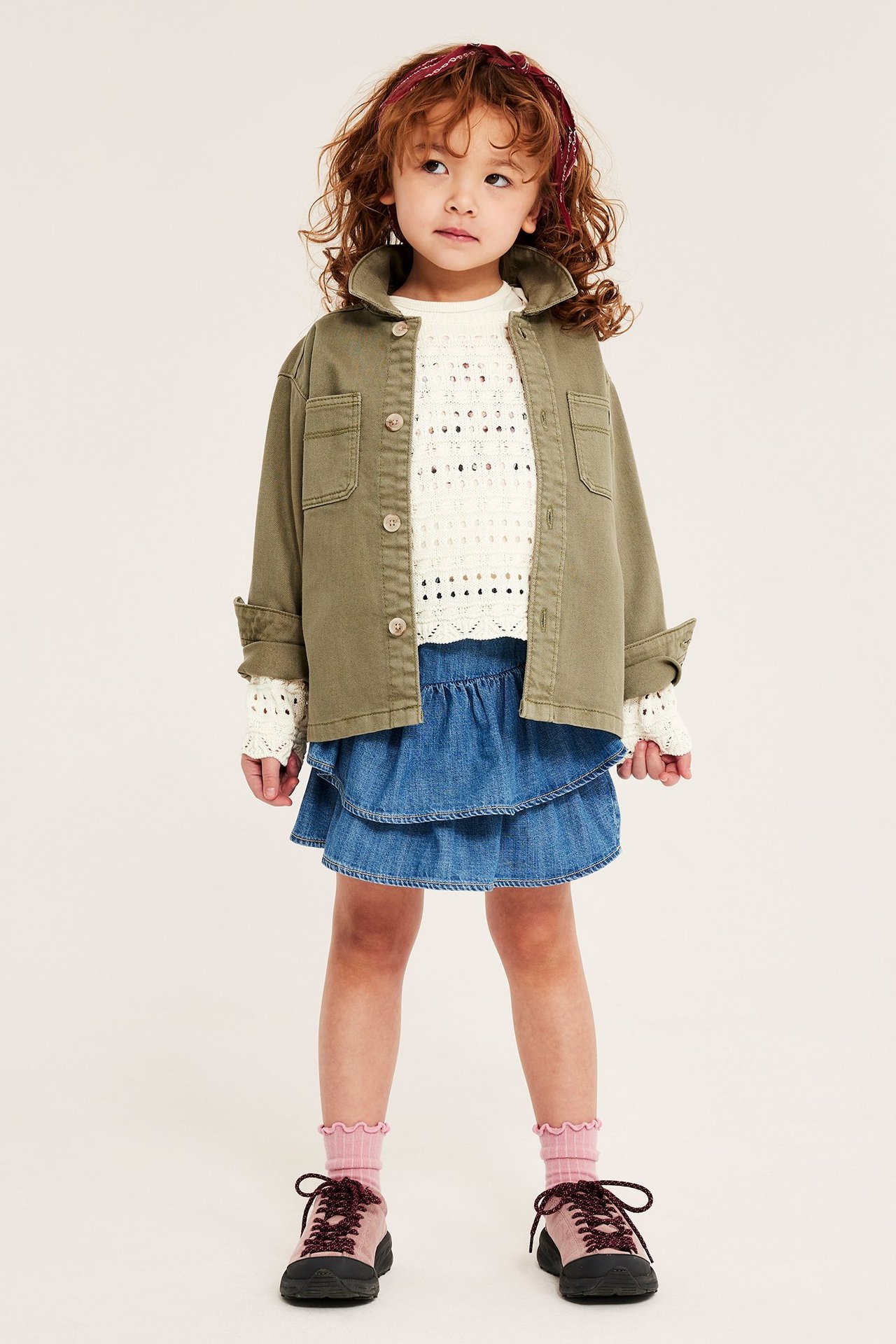 Forfra: Jente i offwhite hullstrikket bomullsgenser, khaki jakke, blått denimskjørt og rosa joggesko.