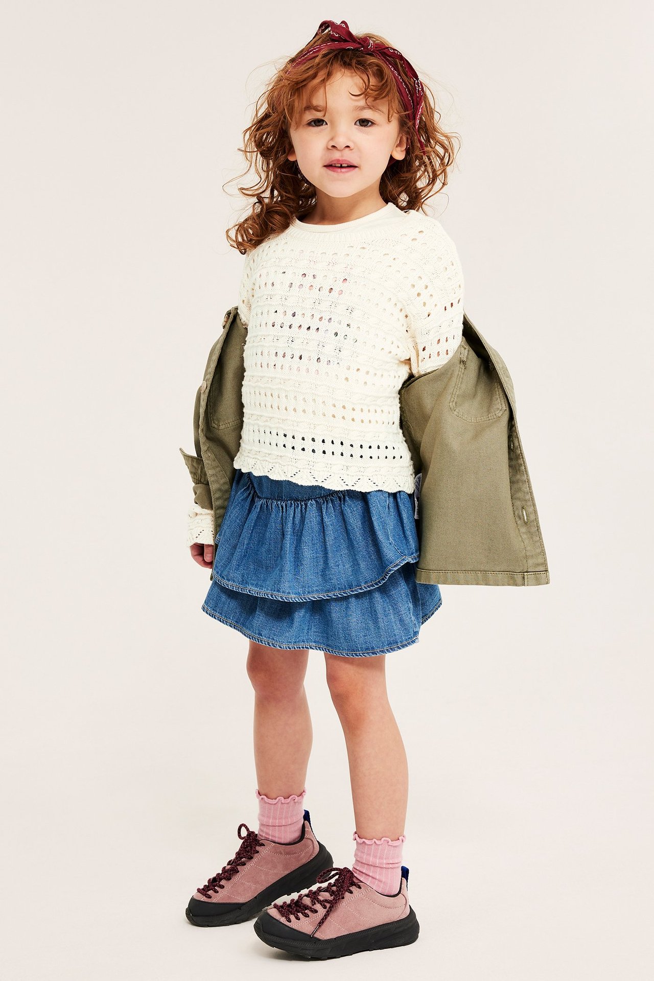 Frontbilde: Barn i offwhite hullstrikket bomullsgenser, blått denimskjørt, oliven jakke, rosa sokker, sneakers.