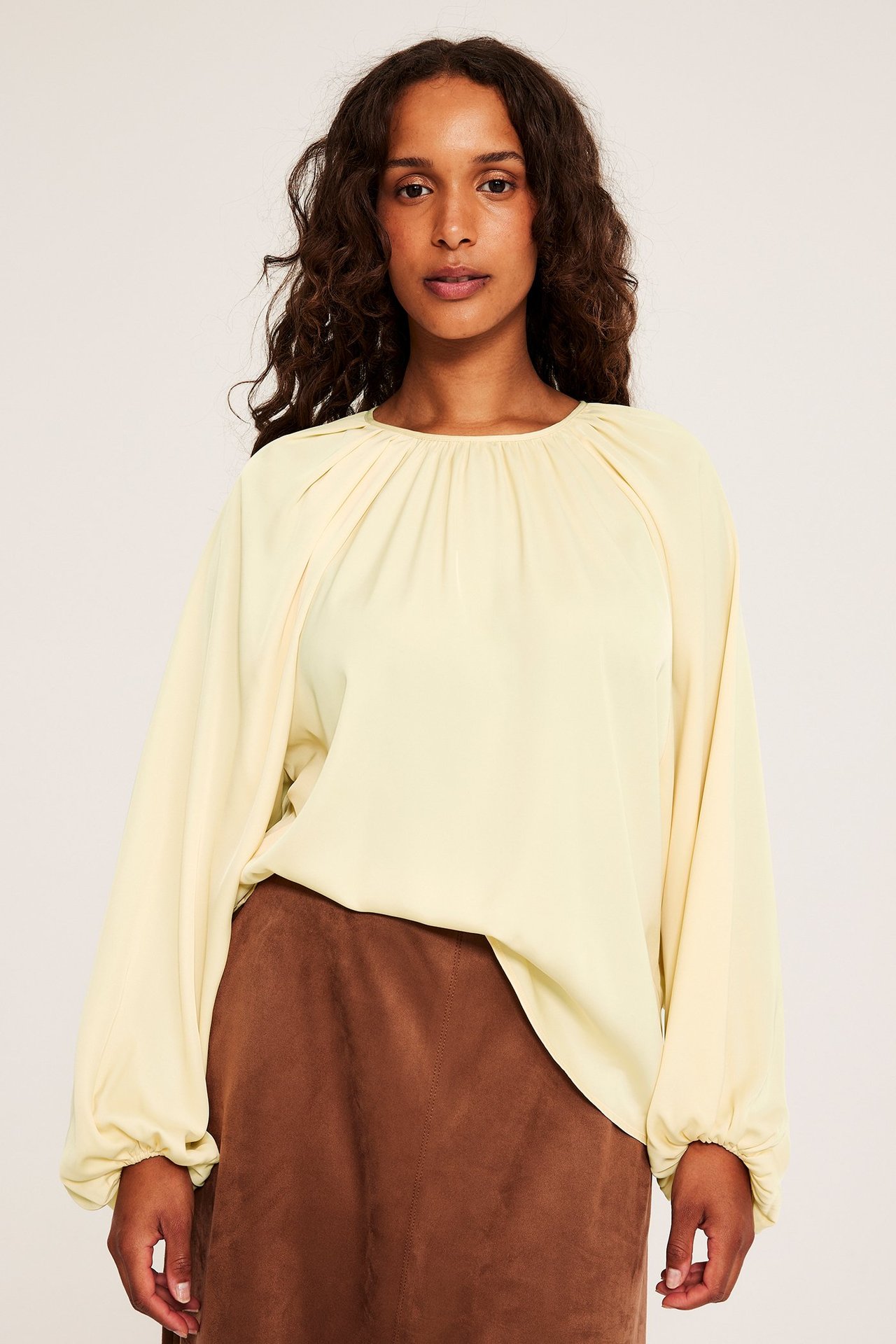 Kvinne i lys gul polyesterbluse med voluminøse ermer, over et brunt skjørt.
