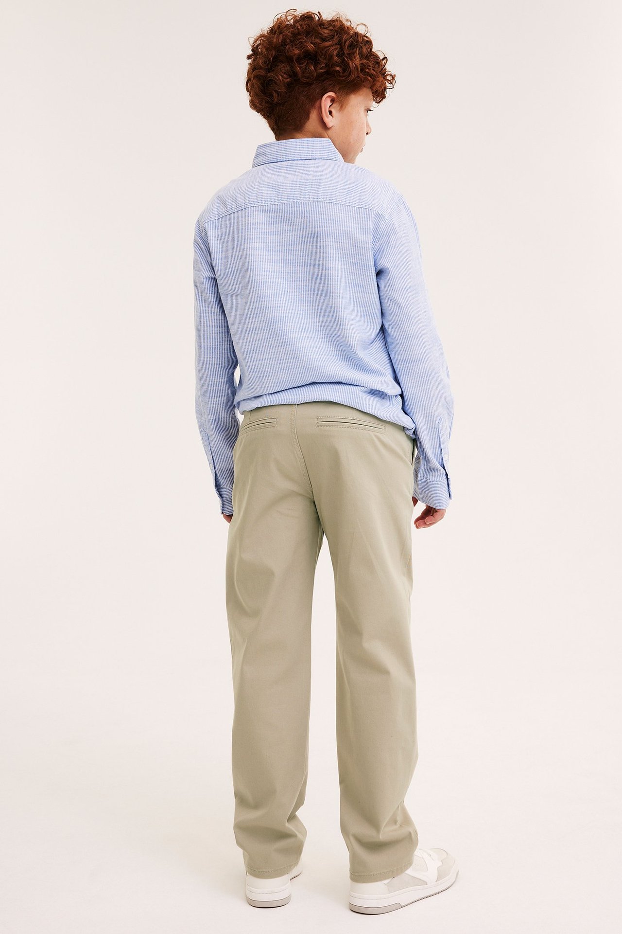 Bakifrån på ett barn i beige stretchchinos med fickor och en ljusblå långärmad skjorta.