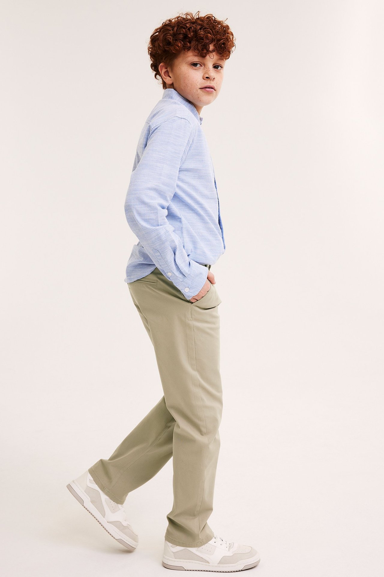 Widok z boku dziecka w beżowych chinosach, jasnoniebieskiej koszuli w paski i białych sneakersach.