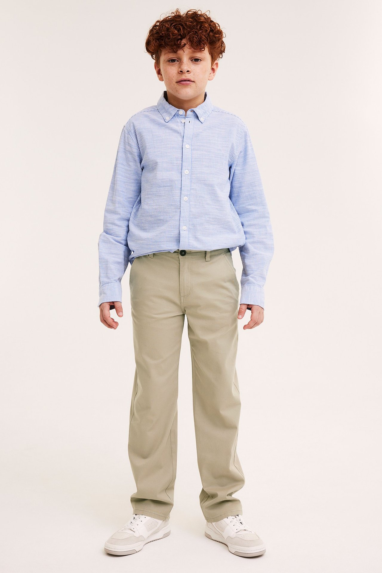 Framifrån, en pojke i beige bomullschinos, en ljusblå randig skjorta och vita sneakers.