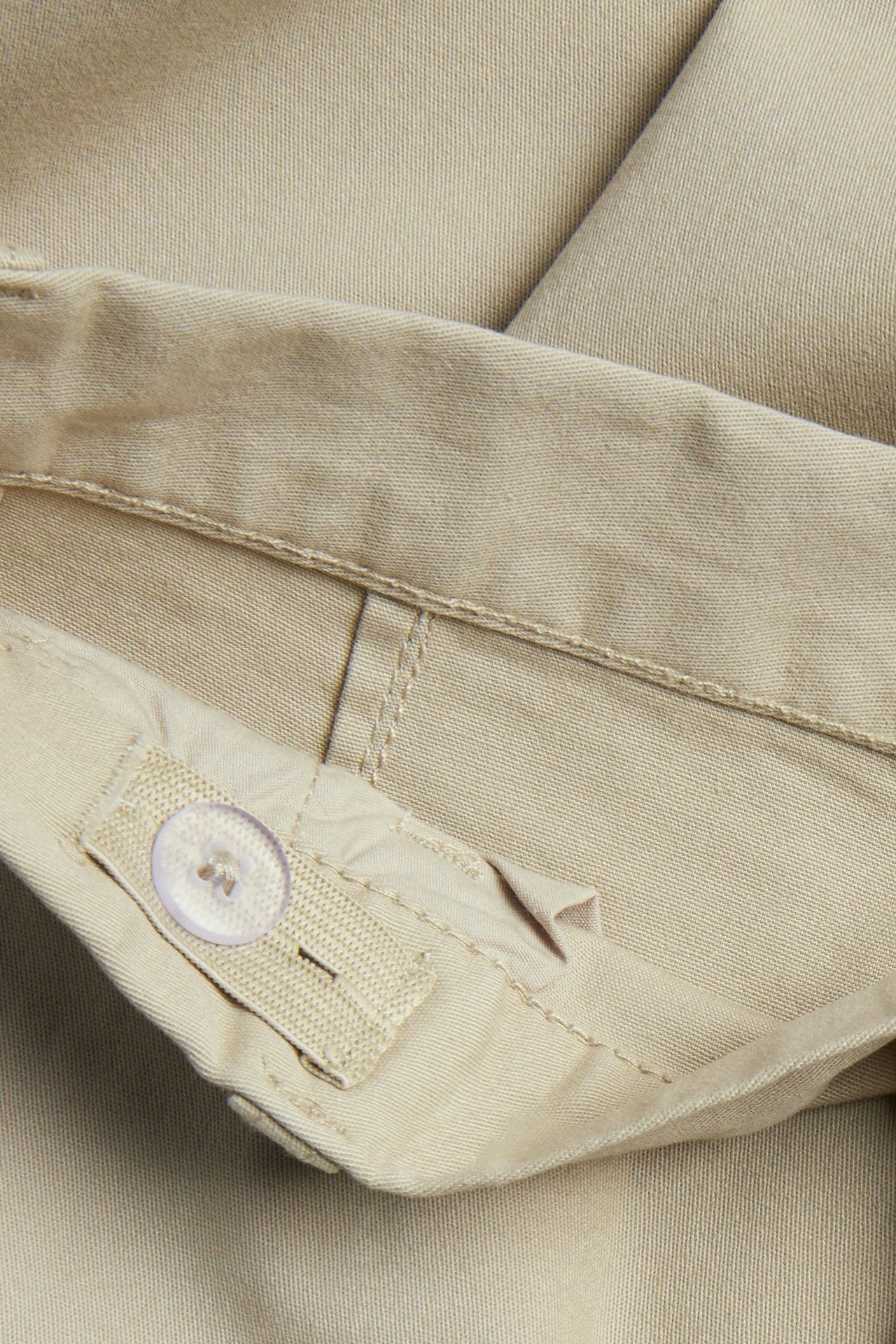 Lähikuva beigestä stretch-puuvillachinoksesta, jossa säädettävä kuminauhavyötärö ja läpinäkyvä nappi.