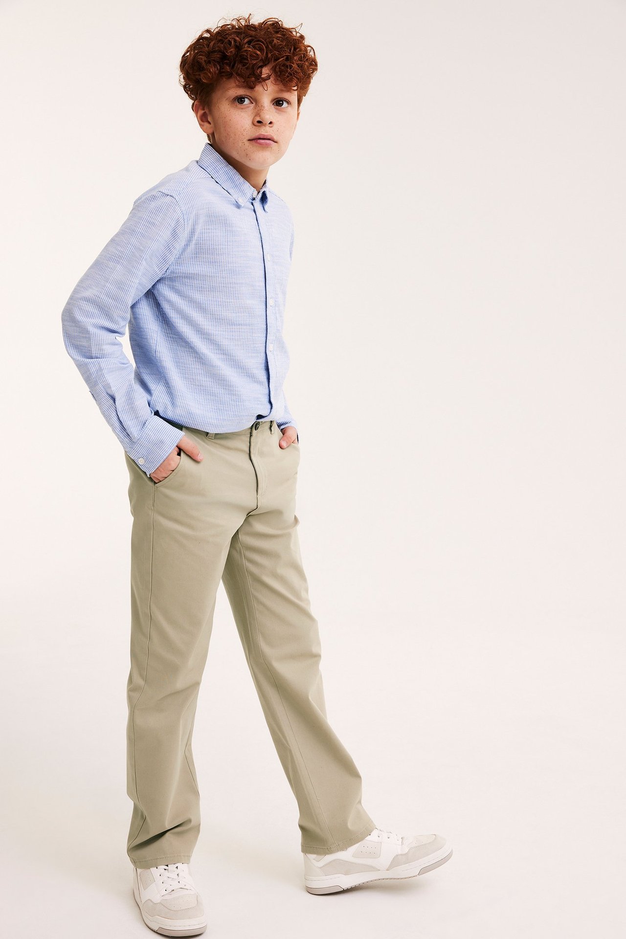 Dziecko w beżowych chinosach stretch, jasnoniebieskiej kraciastej koszuli i białych sneakersach.