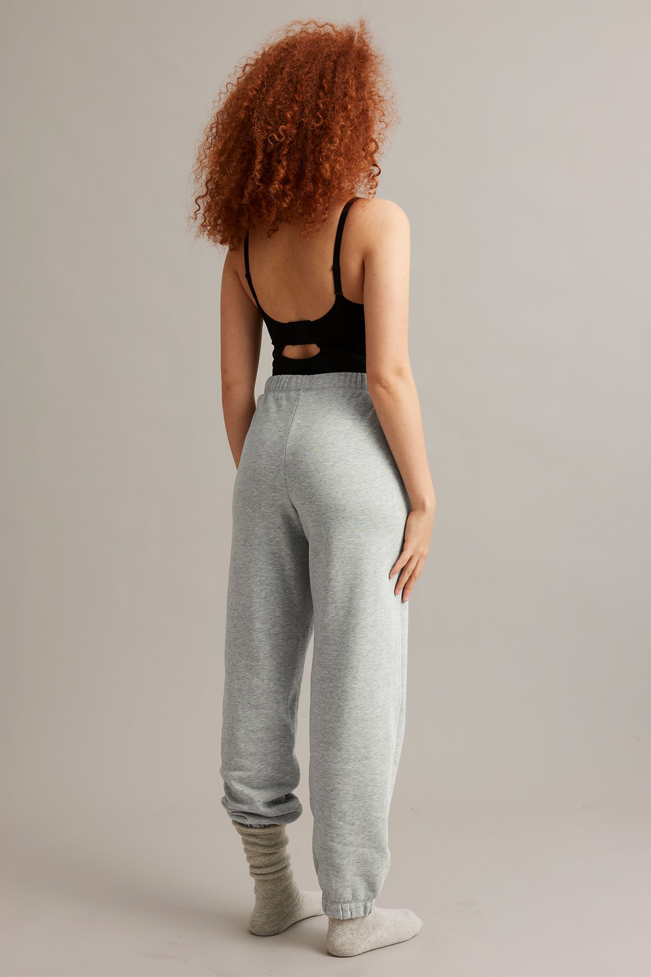 Bakifrån, kvinna i ljusgråmelerade joggers med resårmidja, svart linne och ljusbeige strumpor.