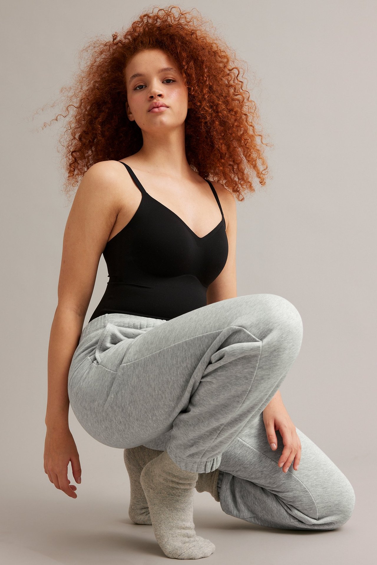 Edestä: Nainen yllään vaaleanharmaat meleeratut collegehousut sivutaskuilla, musta v-toppi ja harmaat sukat.