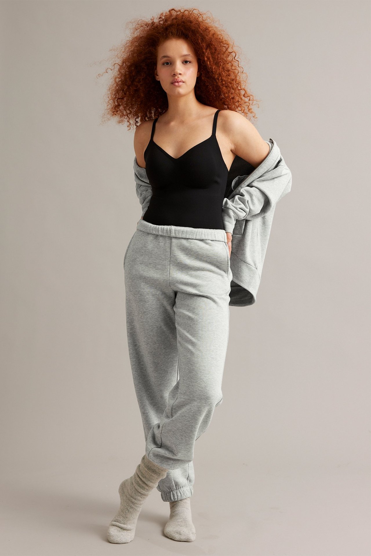 Edestä: Nainen yllään vaaleanharmaat meleeratut collegehousut joustovyötäröllä, musta toppi, harmaa huppari ja sukat.