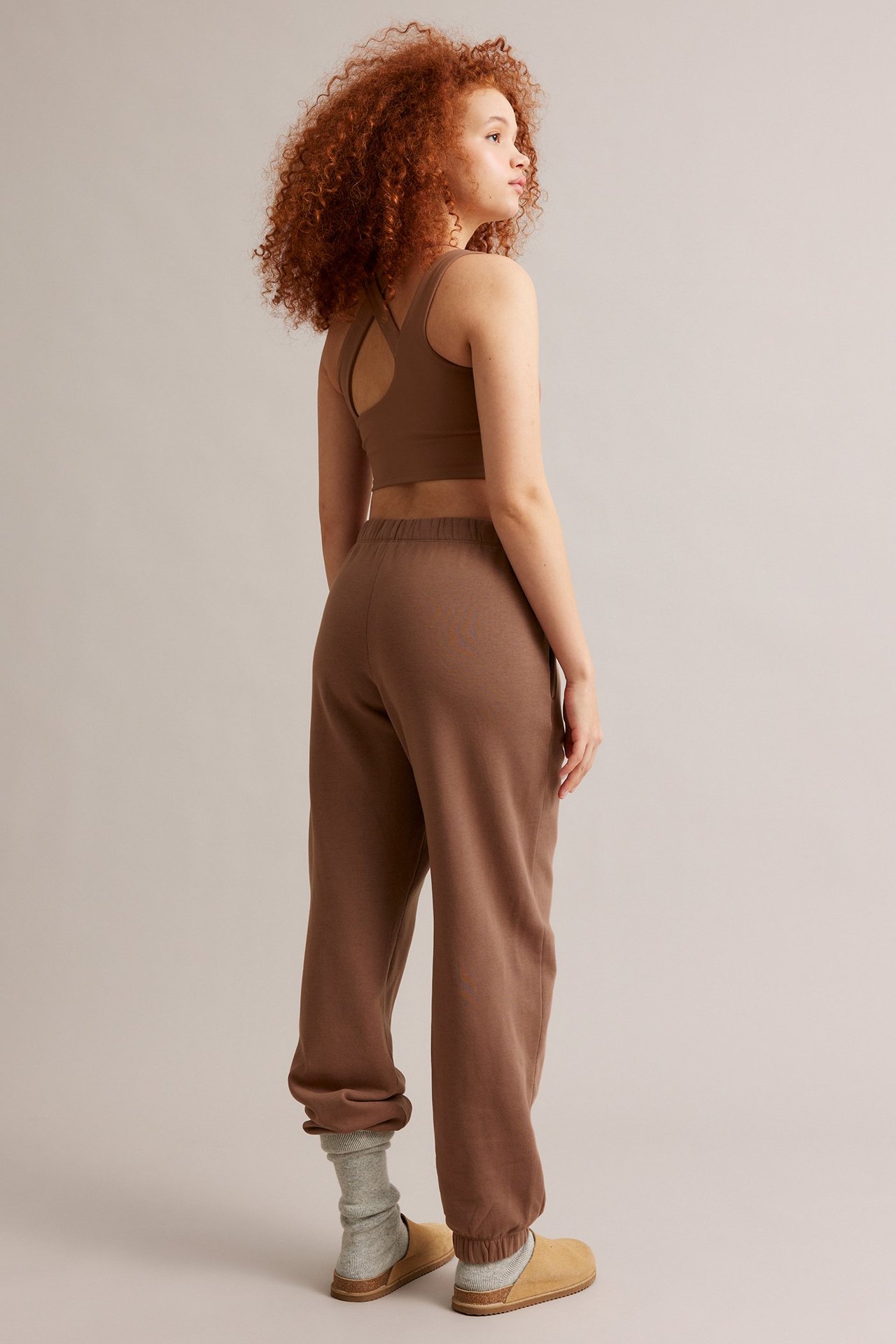 Naisen takasivuprofiili ruskeissa collegehousuissa, yhteensopivissa urheiluliiveissä, vaaleanharmaissa sukissa ja vaaleanruskeissa pistokkaissa.