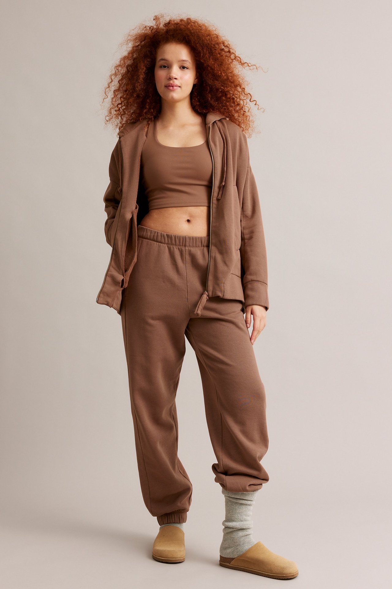 Frontutsikt: kvinne i brune joggebukser, matchende brun crop top og åpen hettegenser.