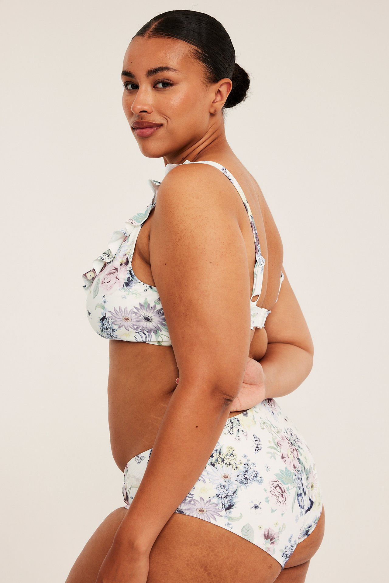 Sidovy, kvinnlig modell i ett vitt blommigt bikini-set med volanger.