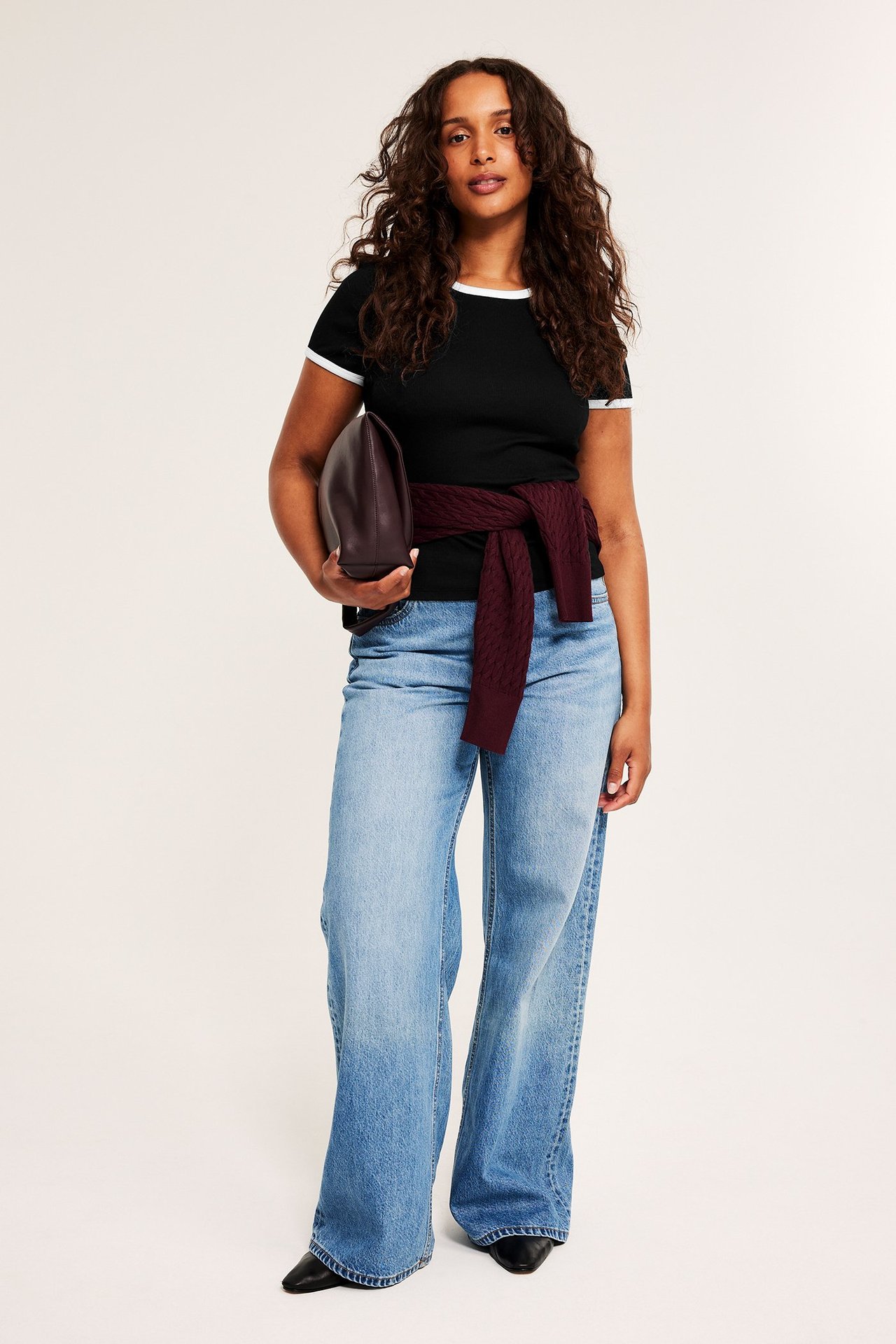 Kobieta w czarnym prążkowanym t-shircie z białym obszyciem, jasnoniebieskich jeansach, trzyma brązową kopertówkę.