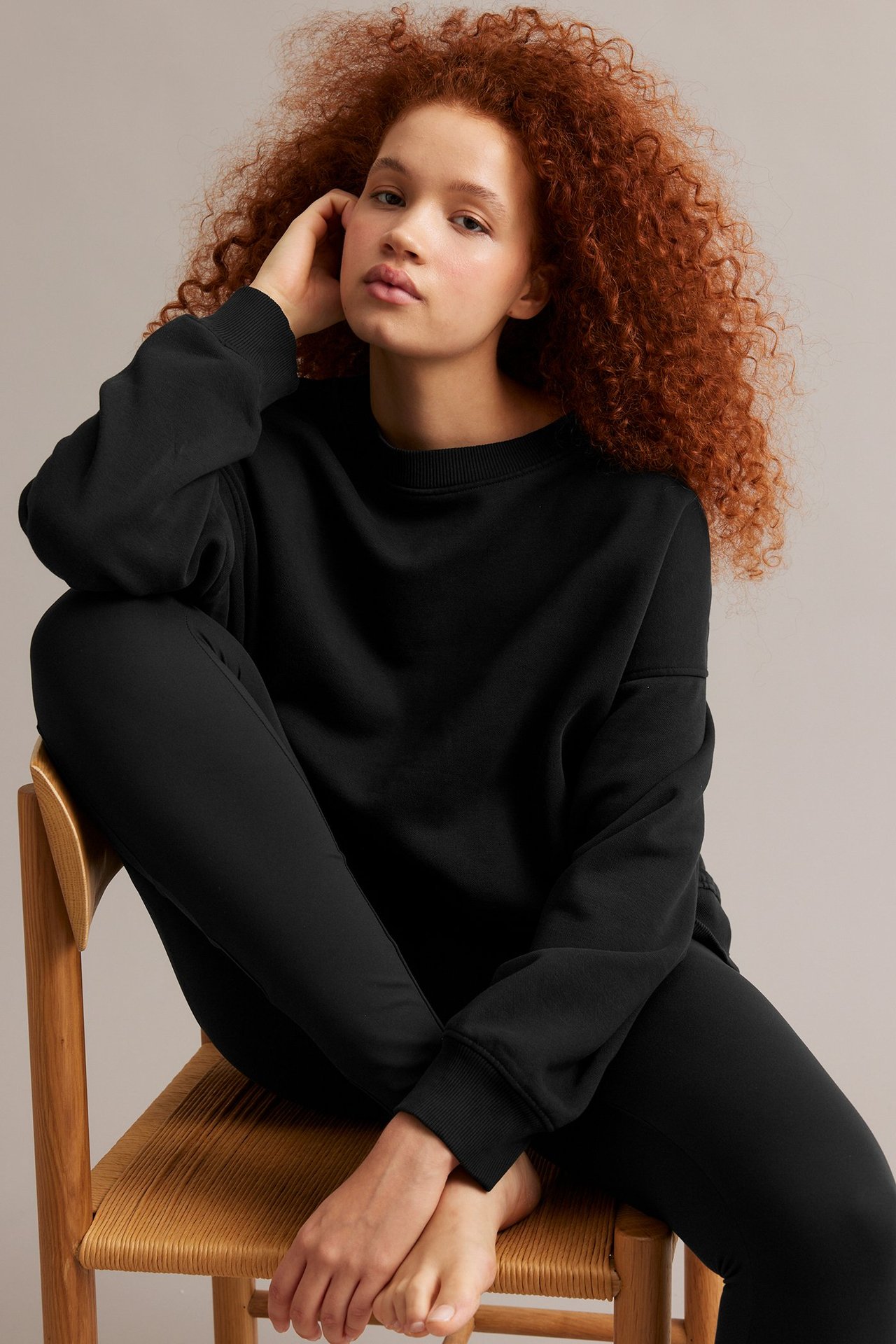 Naisen etukuva mustassa pitkässä collegepaidassa ja mustissa housuissa, istuen paljain jaloin puutuolilla.
