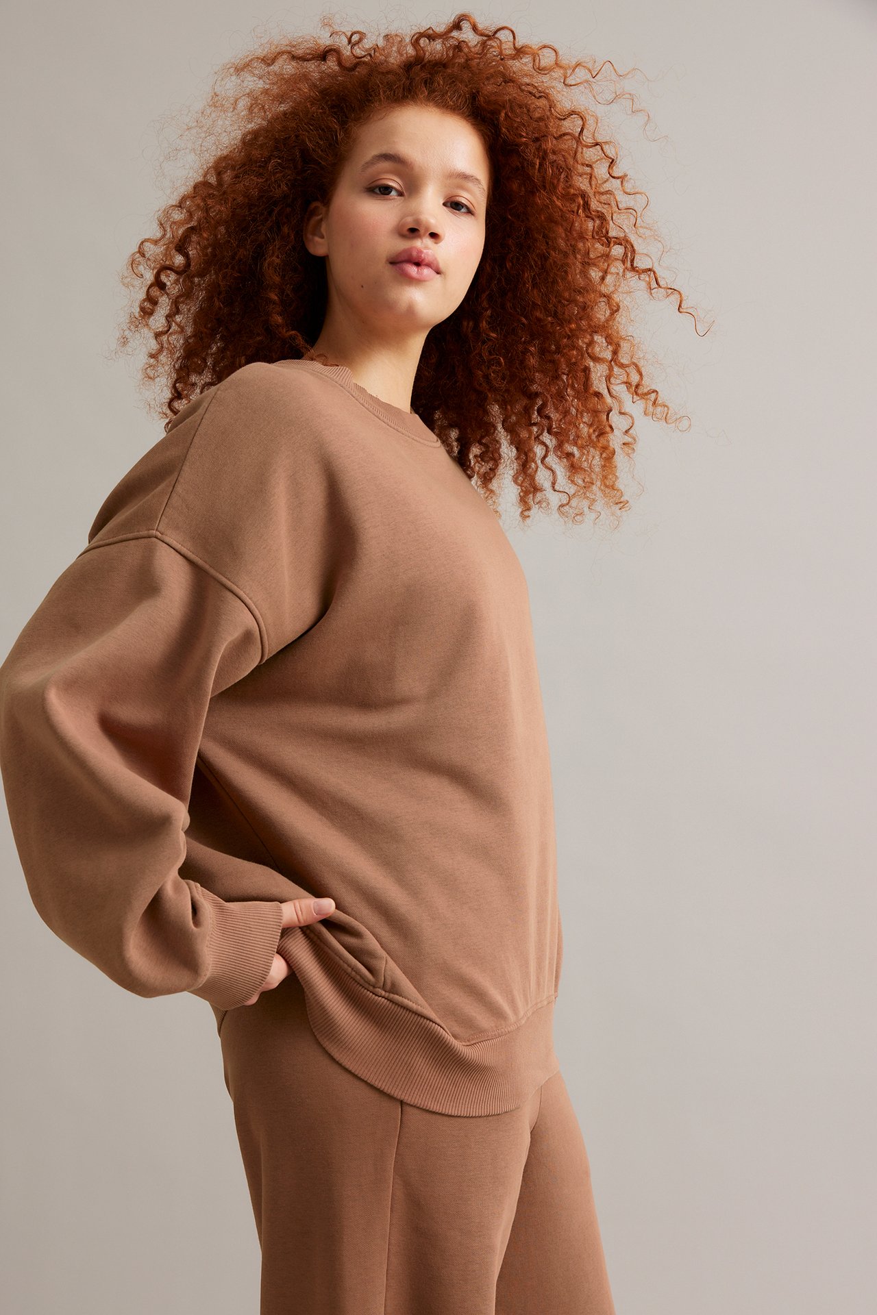 Framifrån, kvinna i lång, lös brun sweatshirt med tvättad look och matchande bruna mjukisbyxor.