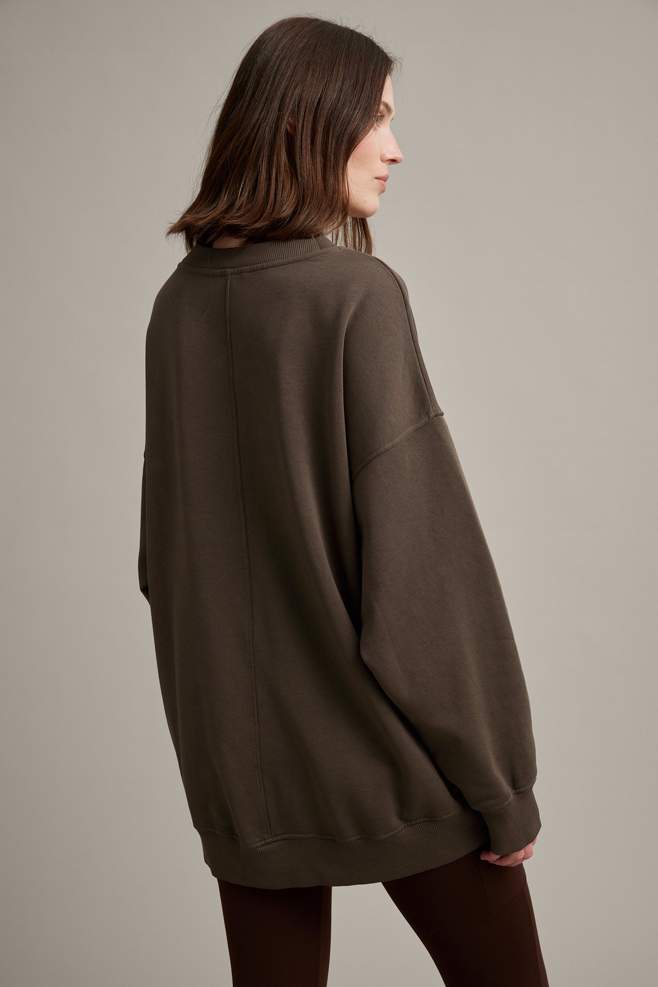 Kvinne, side-bakfra, i oversized mørk brun vasket sweatshirt med mørke leggings.