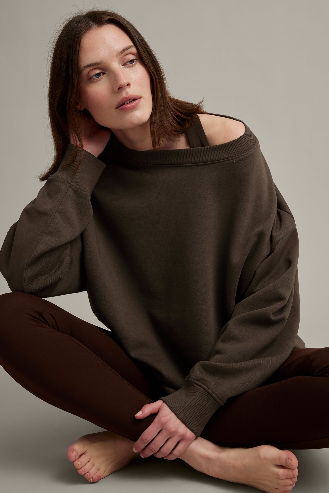 Frontutsikt av en kvinne i en mørkebrun oversized sweatshirt, stylet skulderfri, med mørke leggings.