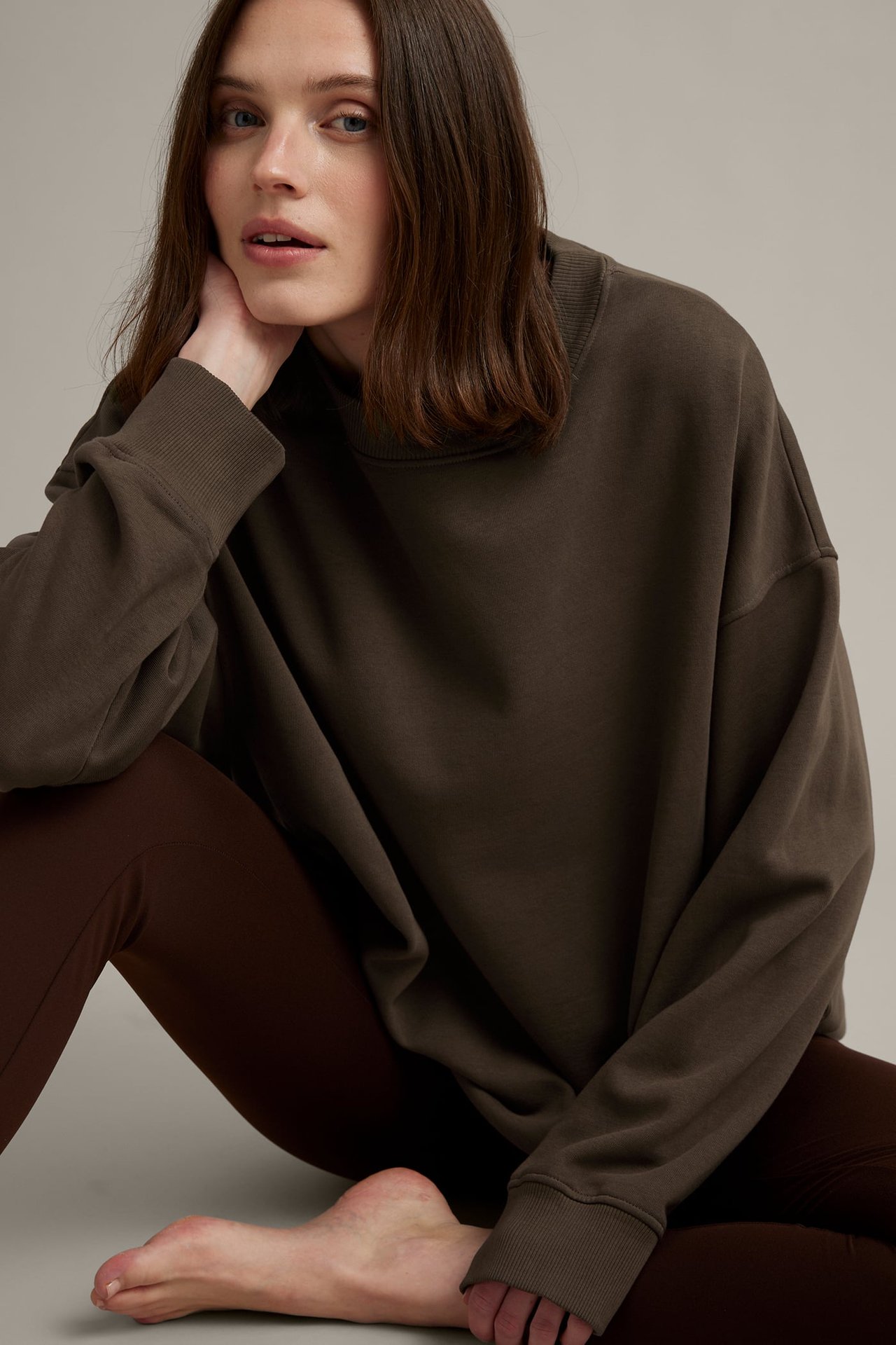 Frontutsikt av en sittende kvinne i mørk brun oversized sweatshirt med ribbekanter og brune leggings.
