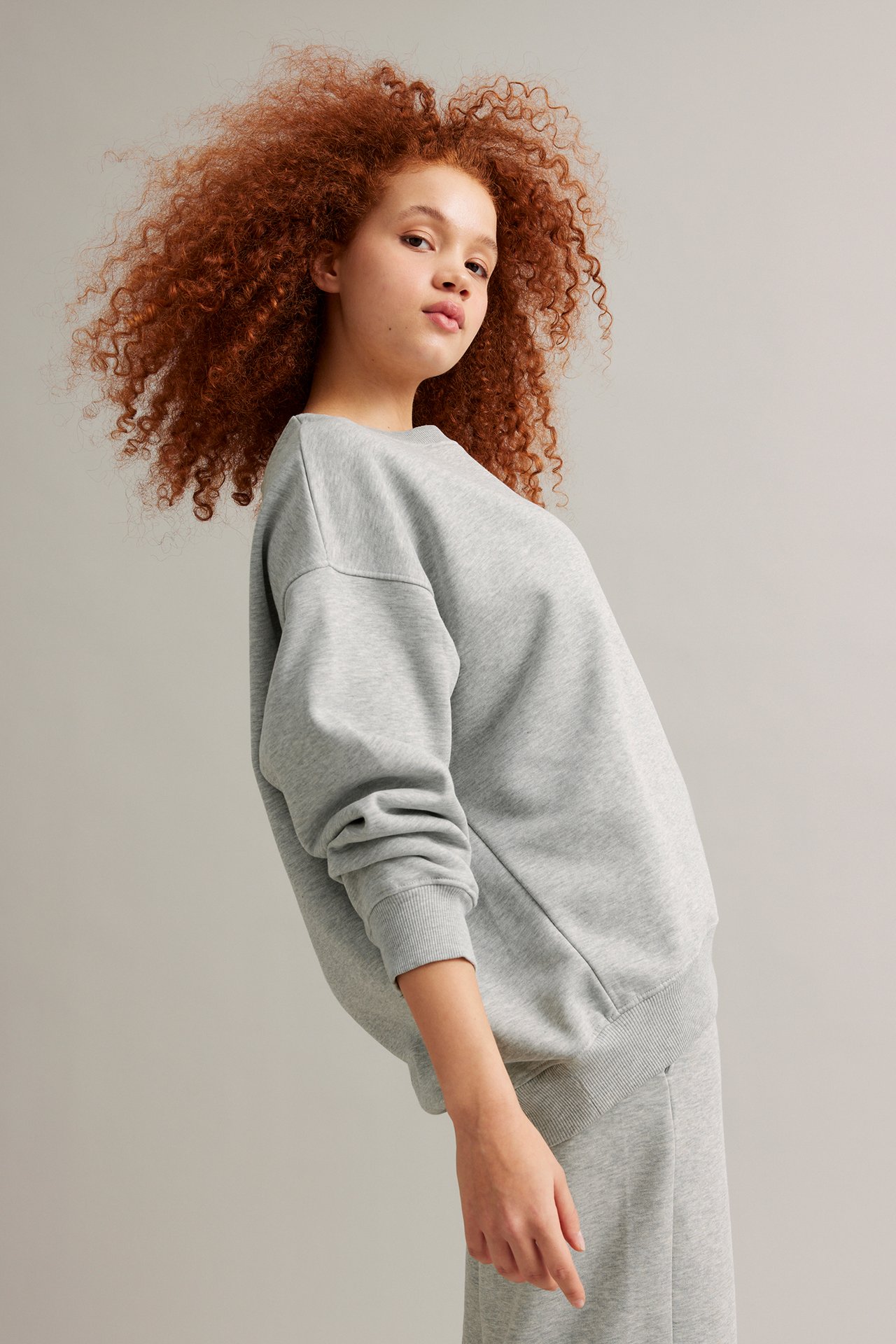 Framifrån: kvinna i lång, lös ljusgrå melerad sweatshirt och matchande sweatpants.