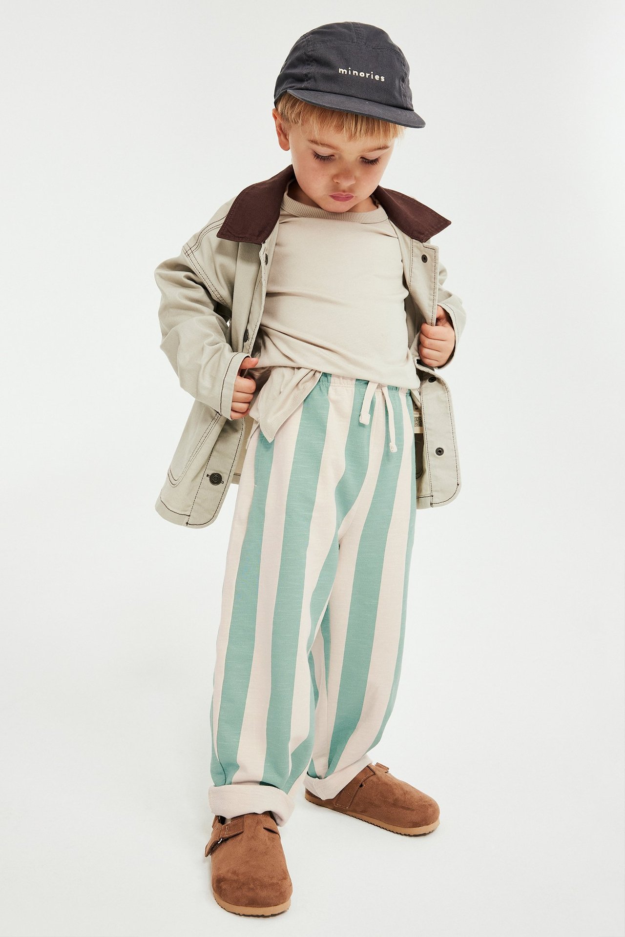 Lapsi yllään minttu/beige raidalliset collegehousut, beige t-paita, vaalea takki, tumma lippalakki, ruskeat kengät.