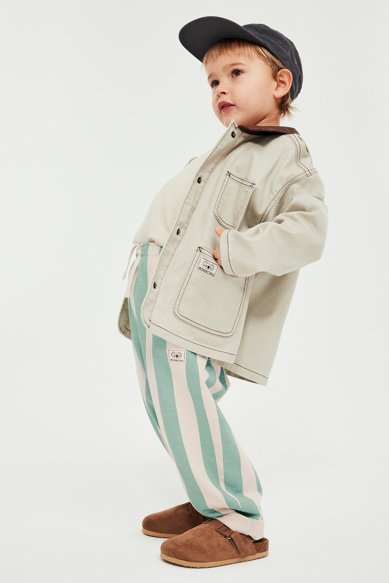 Sivukuva: Lapsi vihreä-valkoraidallisissa collegehousuissa, beigessä takissa, luonnonvalkoisessa t-paidassa, harmaassa lippalakissa ja ruskeissa tohveleissa.