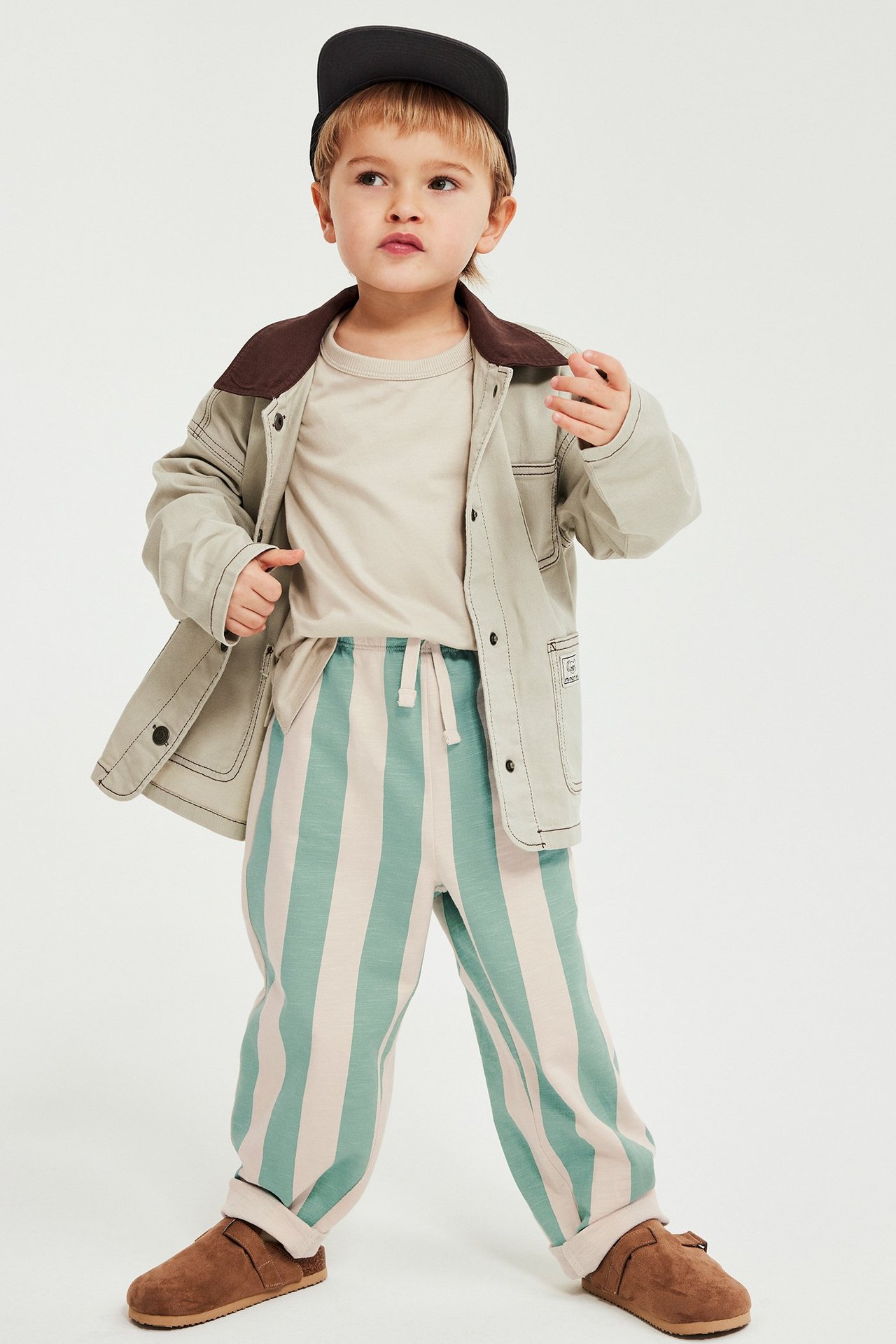 Edestä kuvattu lapsi vihreäraitaisissa collegehousuissa, beigessä t-paidassa, beigessä takissa, mustassa lakissa ja ruskeissa tohveleissa.