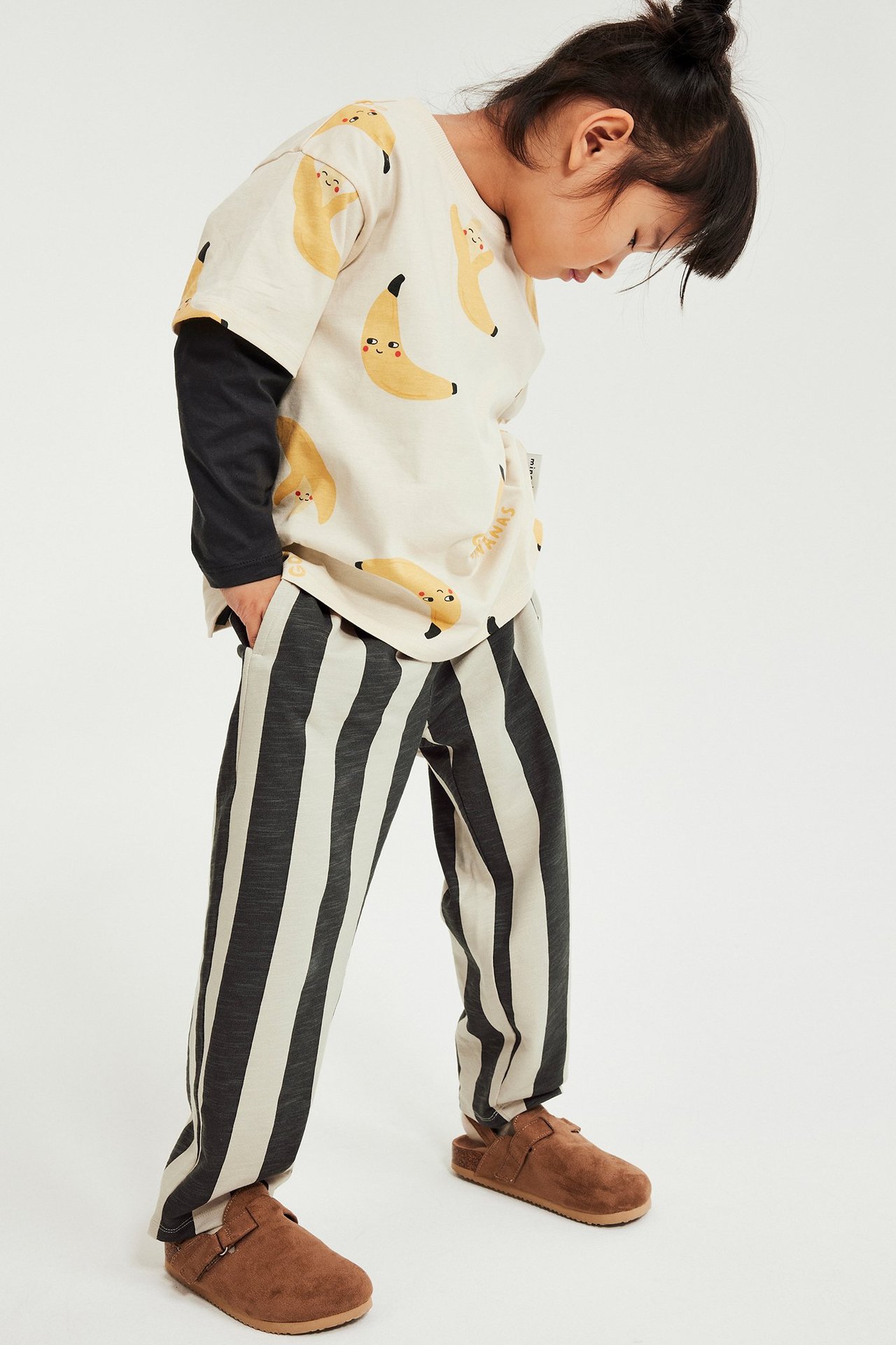 Sidovy av ett barn i svart- och benvita randiga mjukisbyxor, en bananmönstrad t-shirt och bruna träskor.