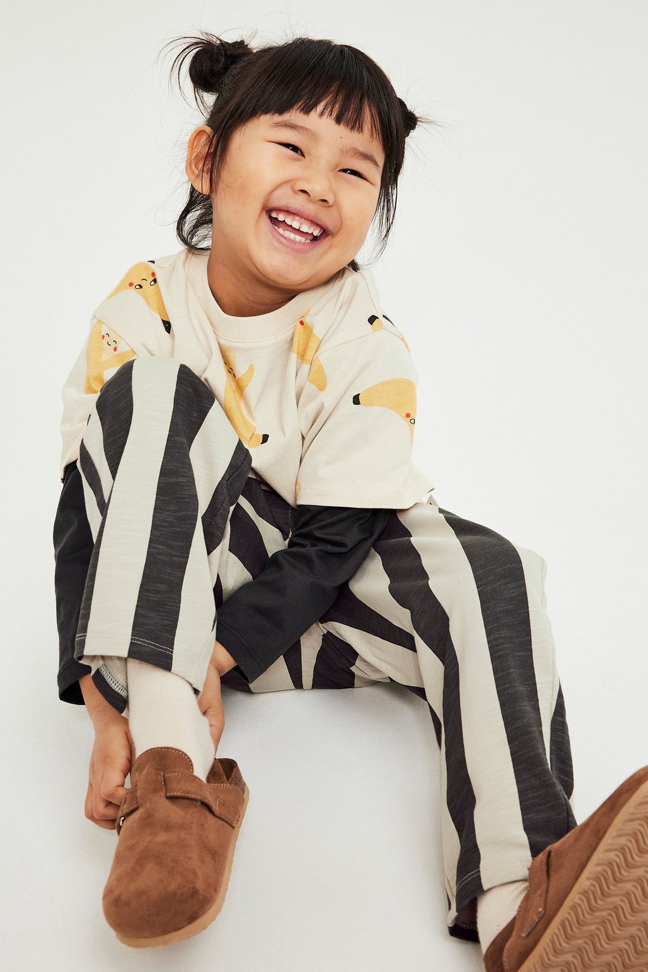 Hymyilevä lapsi yllään musta-beige raidalliset verryttelyhousut, beige eläinkuvioinen t-paita ja ruskeat kengät, edestä.