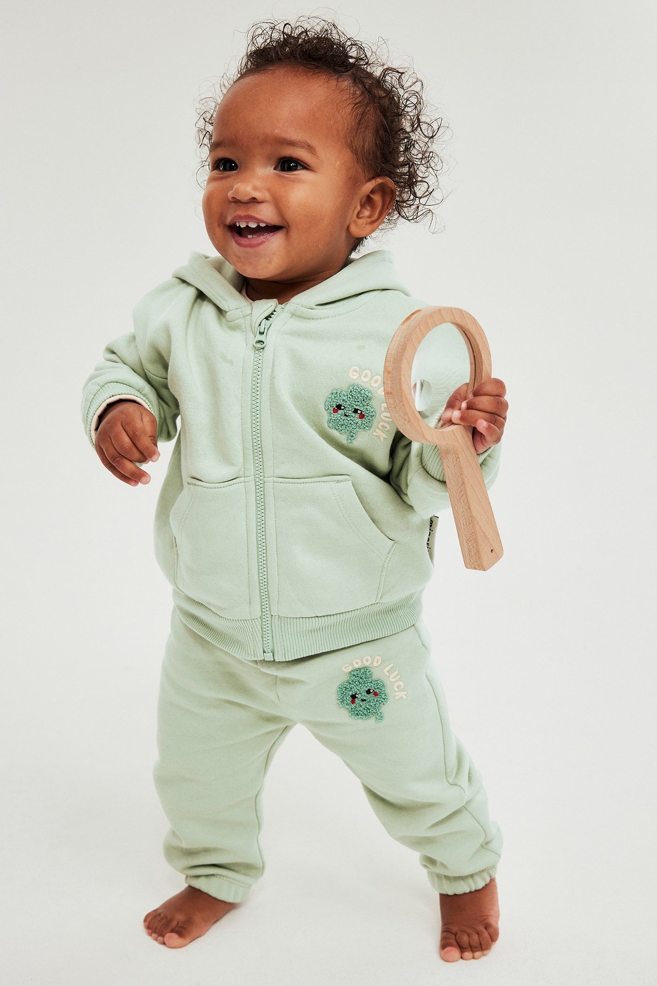 Framifrån: baby i ljusgrön träningsoverall med dragkedja, klöverapplikationer, håller en träspegel.