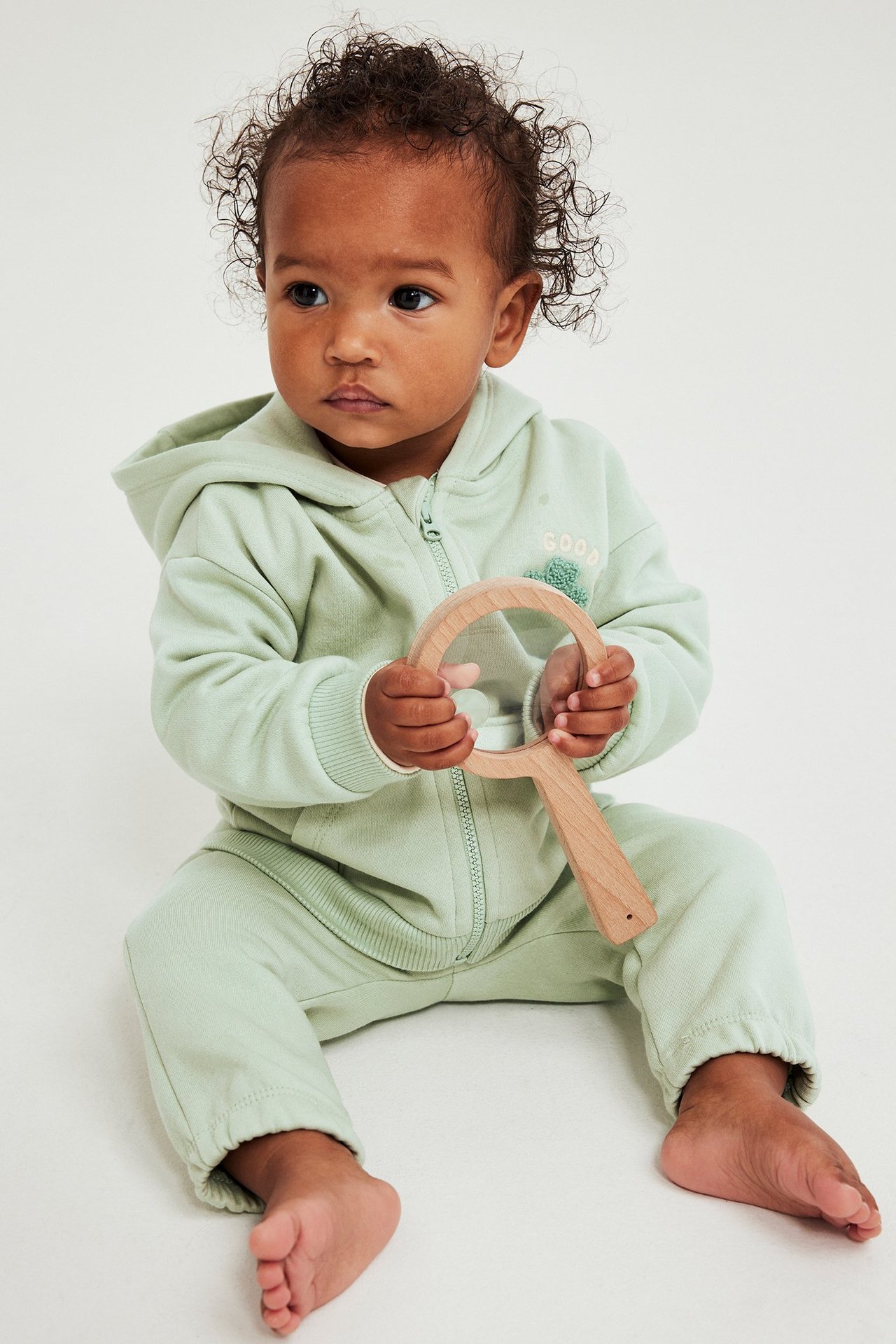 Frontvy av baby i ljusgrön huvtröja och mjukisbyxor med applikation, håller förstoringsglas.